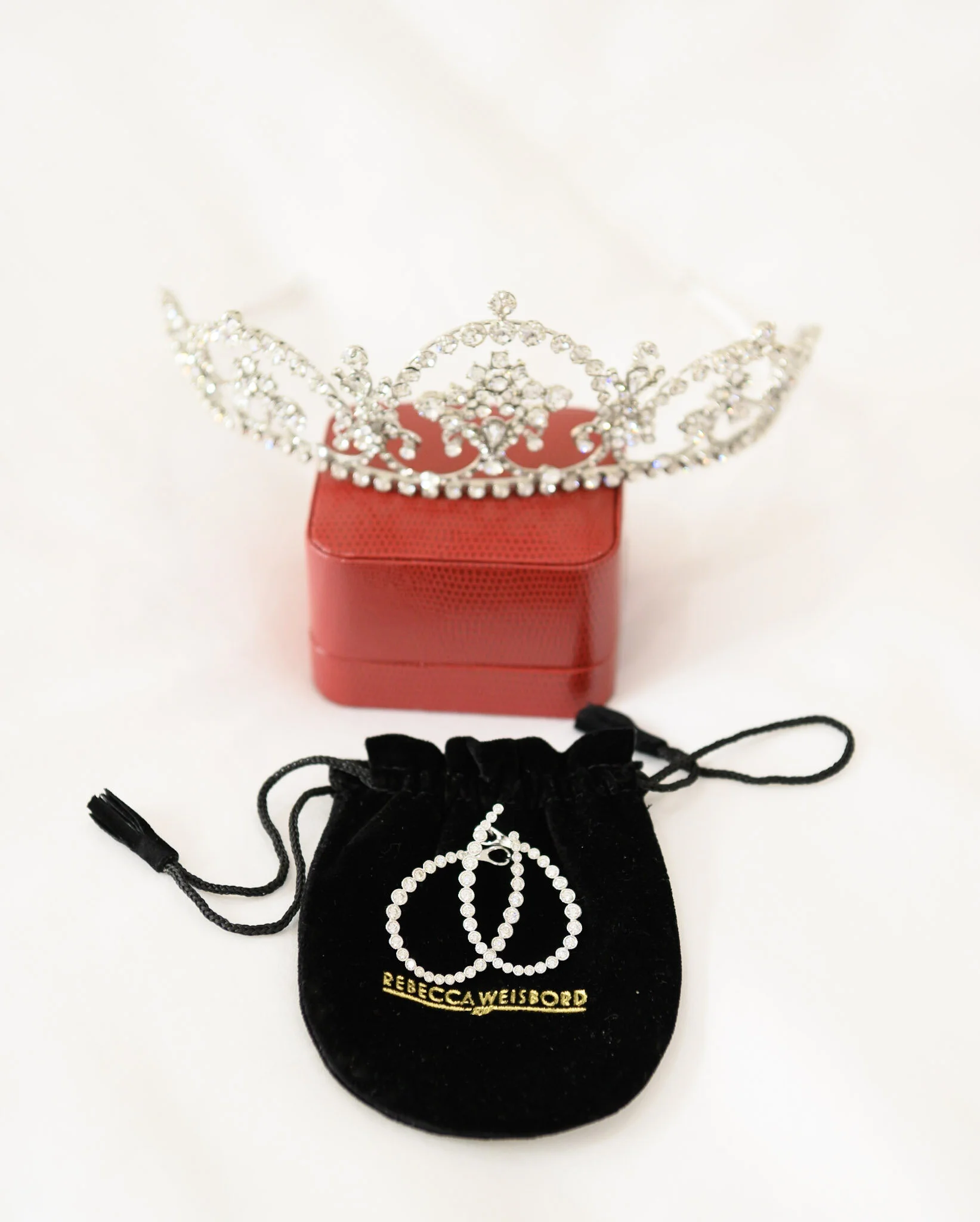 A tiara with rhinestones on a white background, placed on a red box, with a black drawstring bag and jewelry box in front, including a necklace with a peace symbol and the name Rebecca Weisbord.