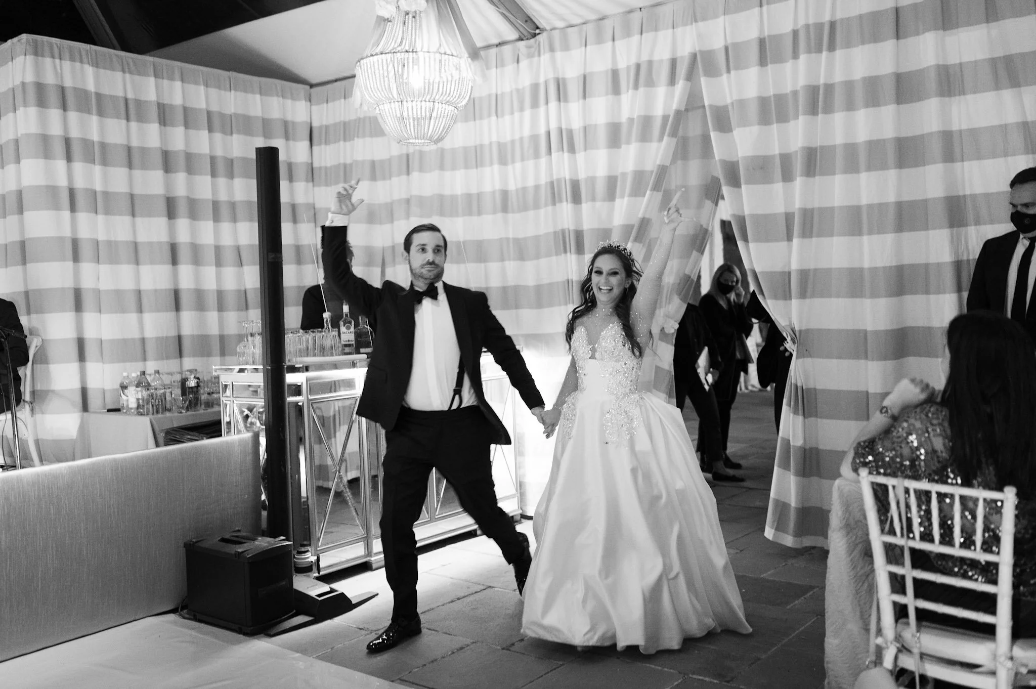 A bride and groom are walking hand in hand at their wedding reception, smiling and celebrating under a striped curtain. The bride is wearing a wedding gown and tiara, while the groom is dressed in a tuxedo. Guests and staff are visible in the backgro