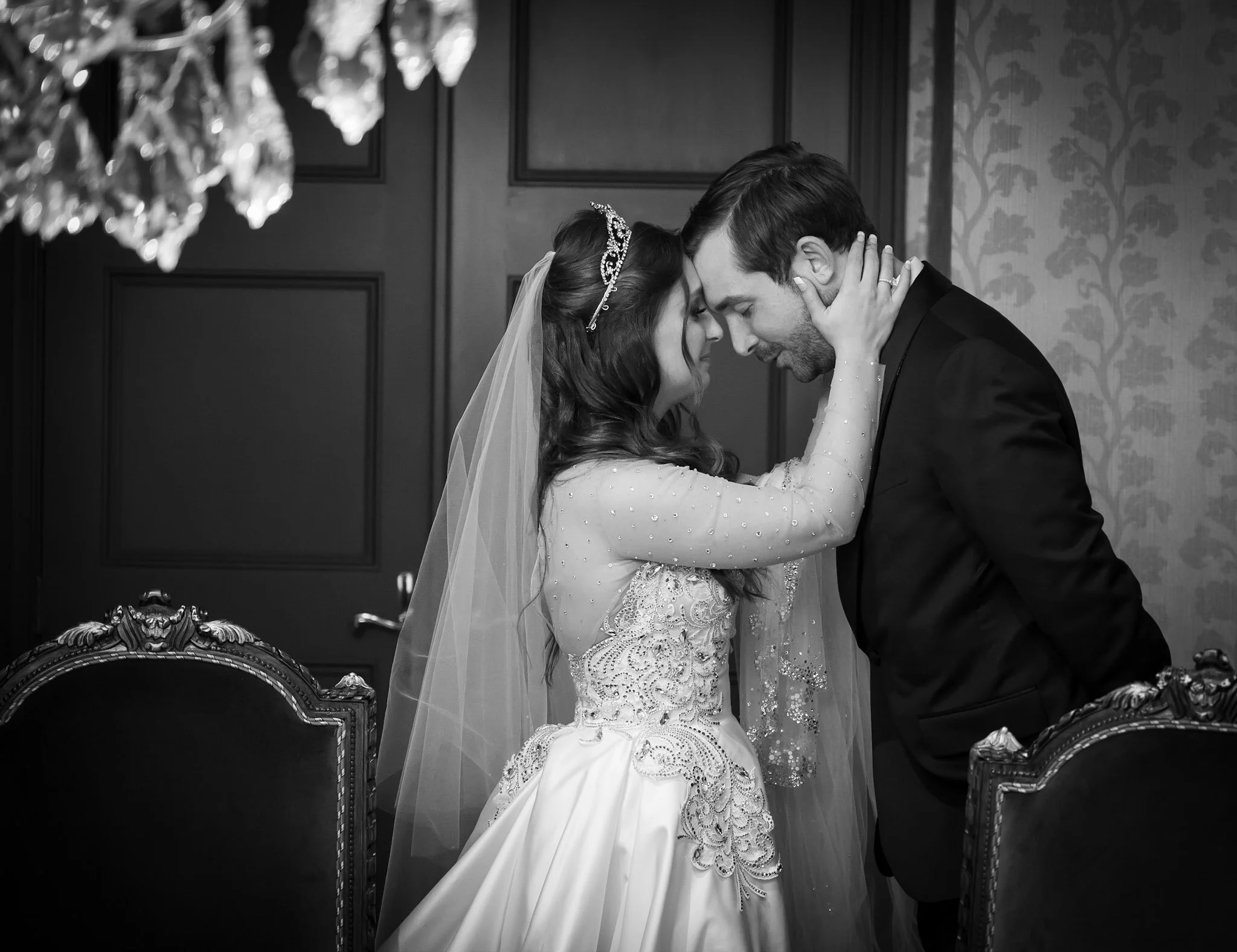 A bride and groom share an intimate moment with foreheads touching and eyes closed inside a decorated room. The bride wears a wedding dress with intricate beading, a veil, and a tiara. The groom wears a dark suit. The background features patterned wa