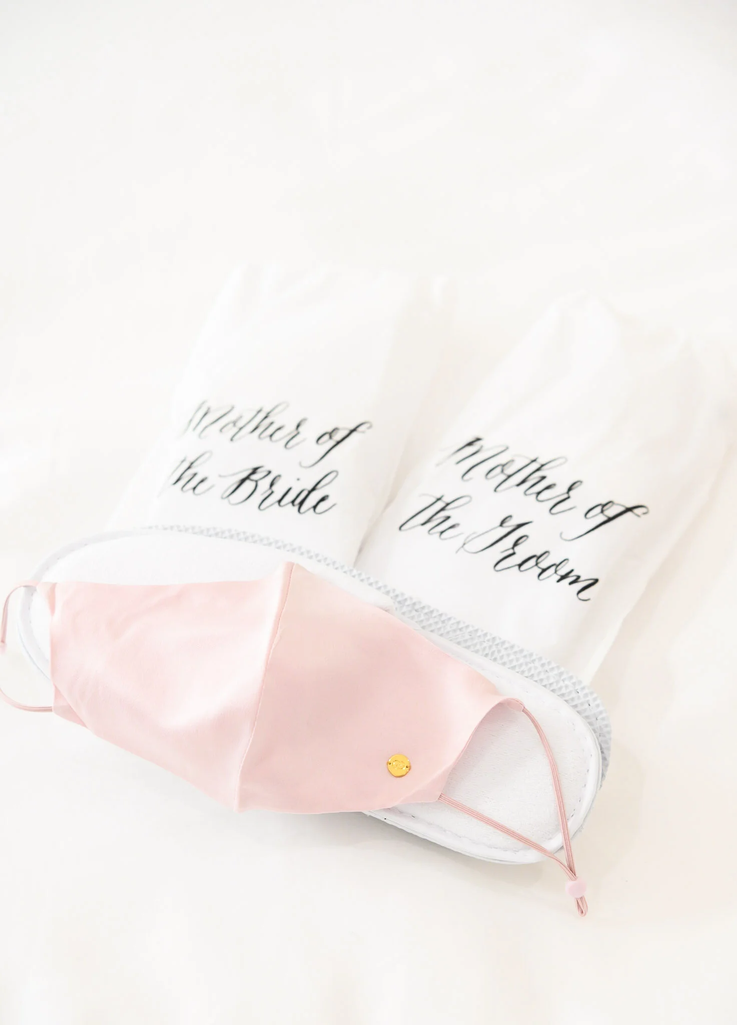 A pink face mask with a gold smiley face button sits on a white surface, partially covering two white paper hand fans with black cursive text reading, 'Mother of the Bride' and 'Mother of the Groom'.