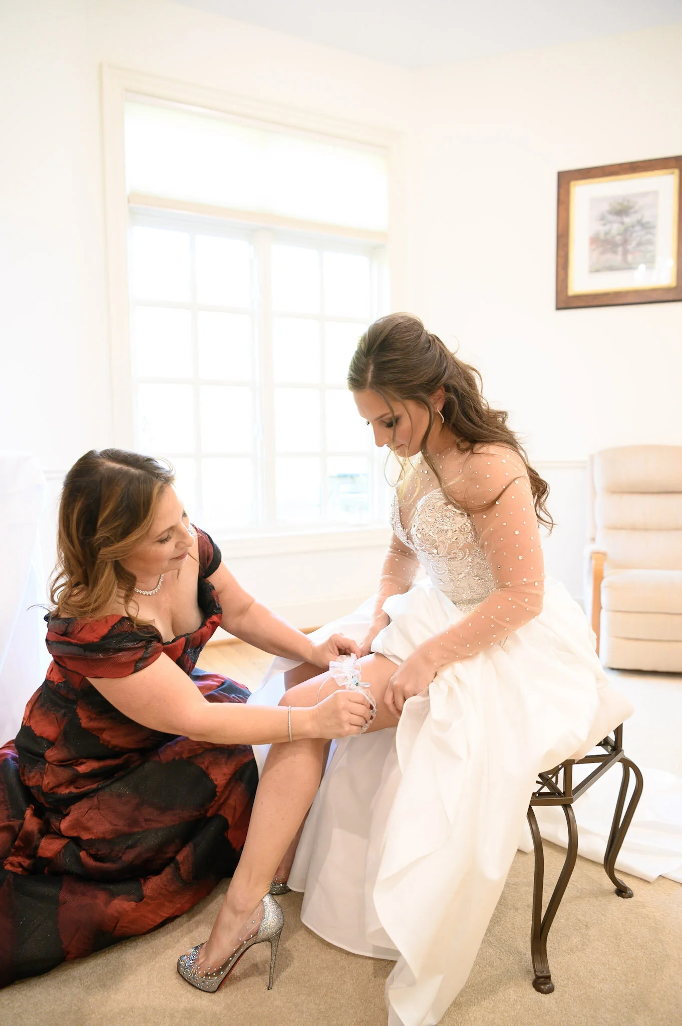 A woman in a wedding dress sitting on a chair with another woman helping her with her shoe, inside a bright room with large windows, wall art, and a beige armchair.