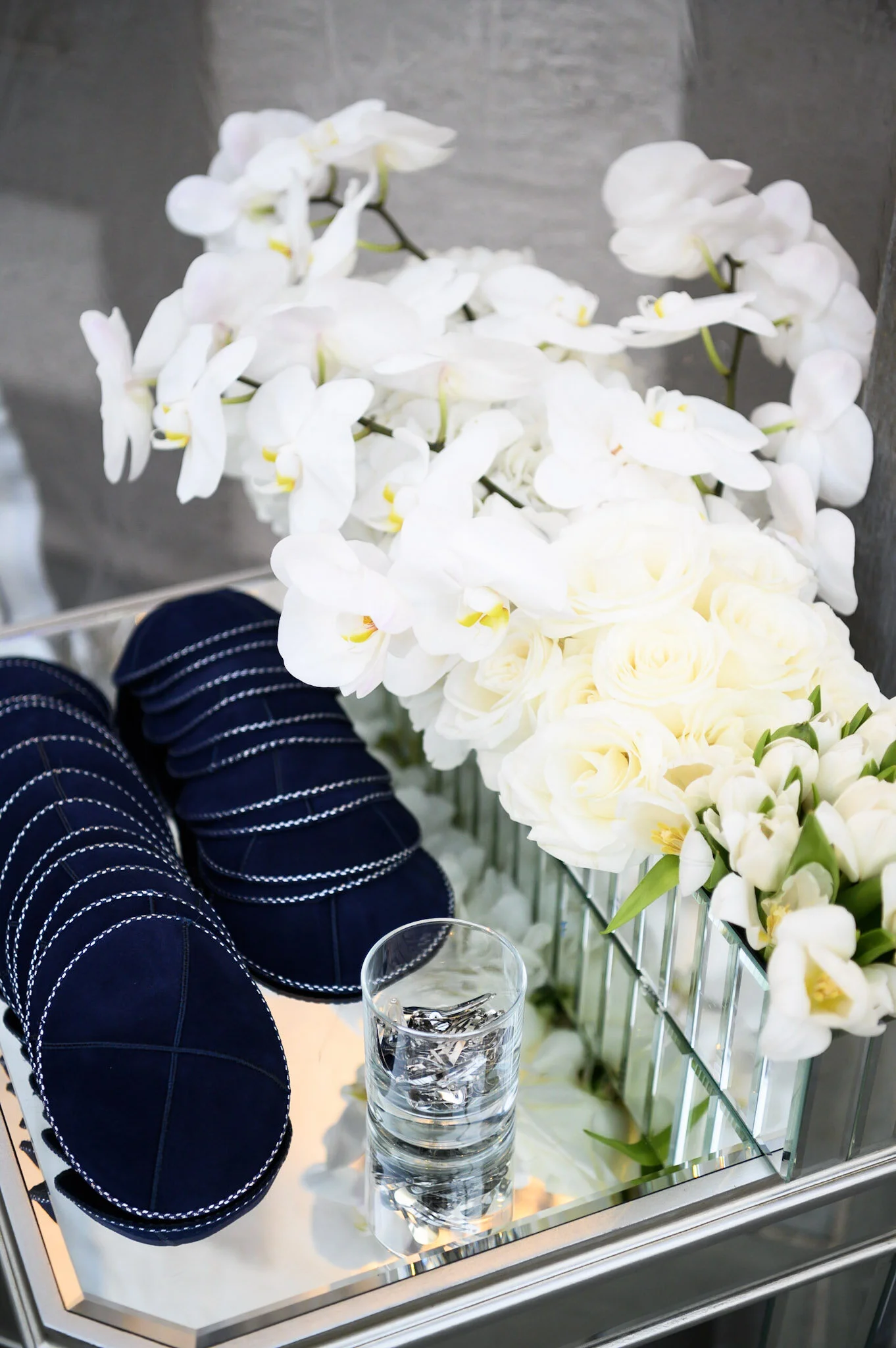 A mirror with a display of white orchids, pale yellow roses, and glass of water on a mirrored surface in a fancy setting.