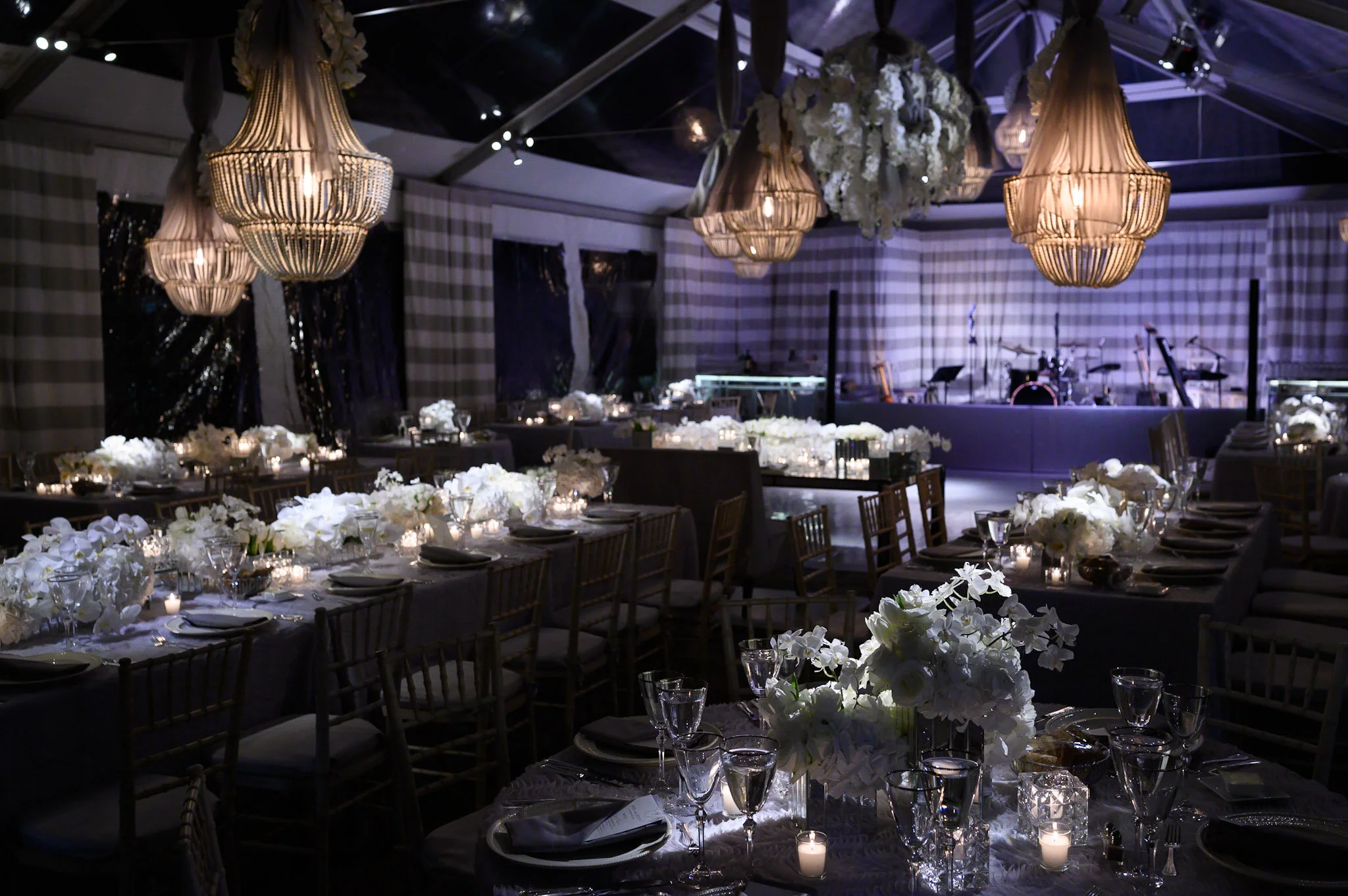 Elegant banquet hall decorated with floral centerpieces and candles, featuring ornate chandeliers hanging from the ceiling and a stage area in the background with musical instruments.