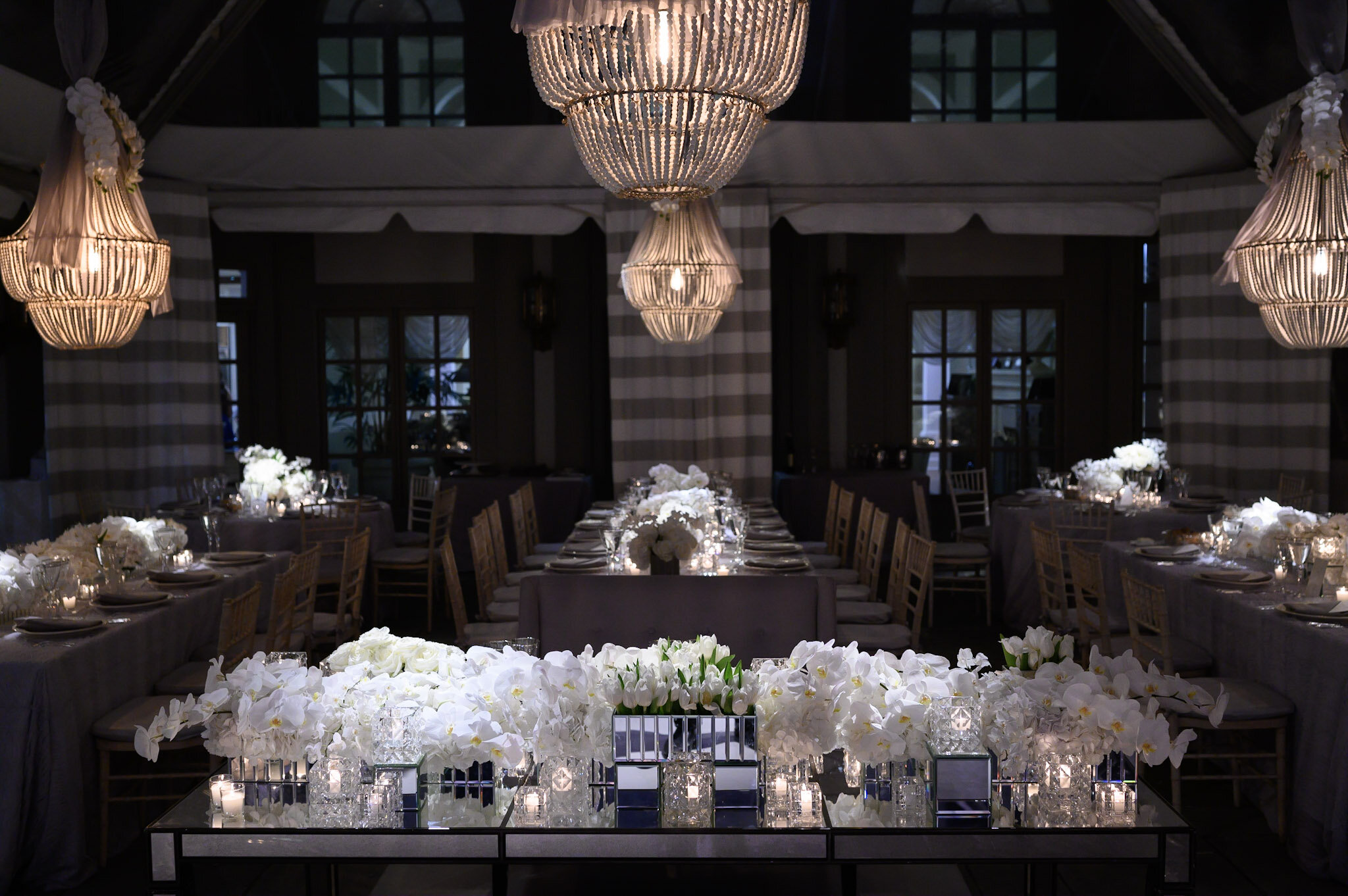 Elegant event space with hanging chandeliers, decorated tables, and floral centerpieces, prepared for a formal celebration or wedding reception.