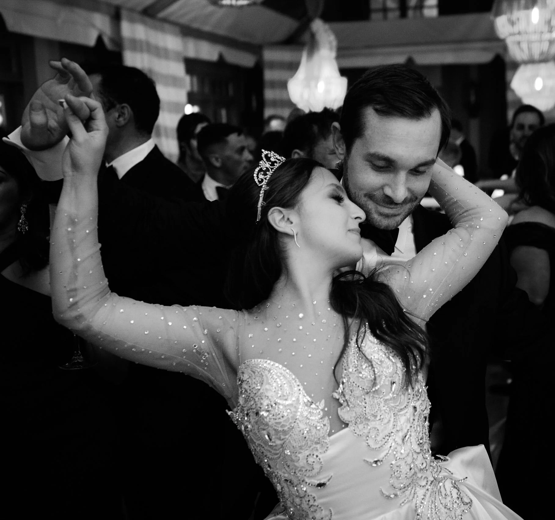 A black and white photo of a couple dancing closely at a formal event, with guests in the background. The woman is wearing an elegant dress with intricate embroidery, and the man is dressed in a suit with a bow tie.