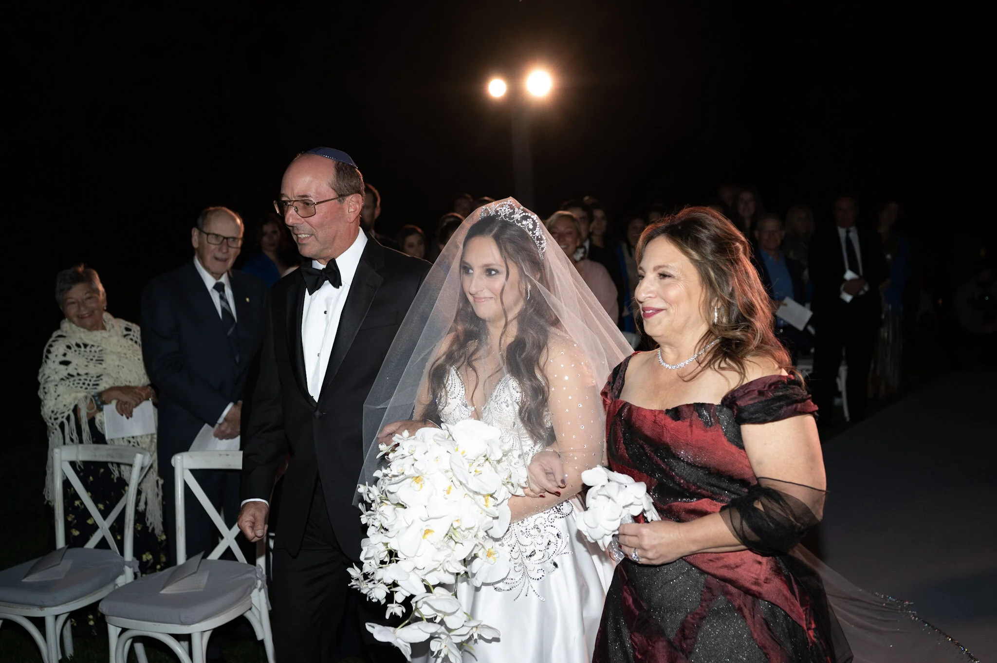 A bride walking down the aisle with her father at a wedding ceremony during nighttime, surrounded by seated guests and illuminated by outdoor lights.