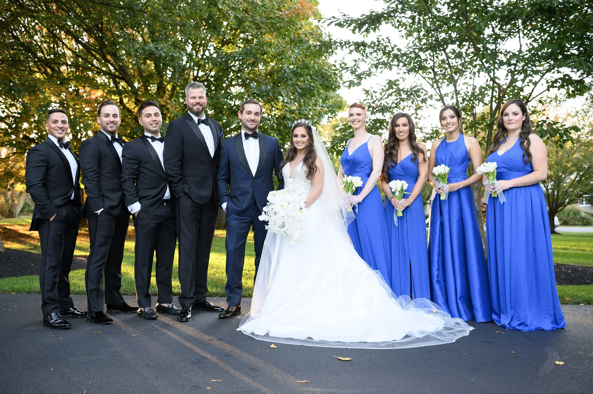 Group of people dressed in formal wedding attire, standing outdoors under trees with green leaves, with some sunlight filtering through the branches.