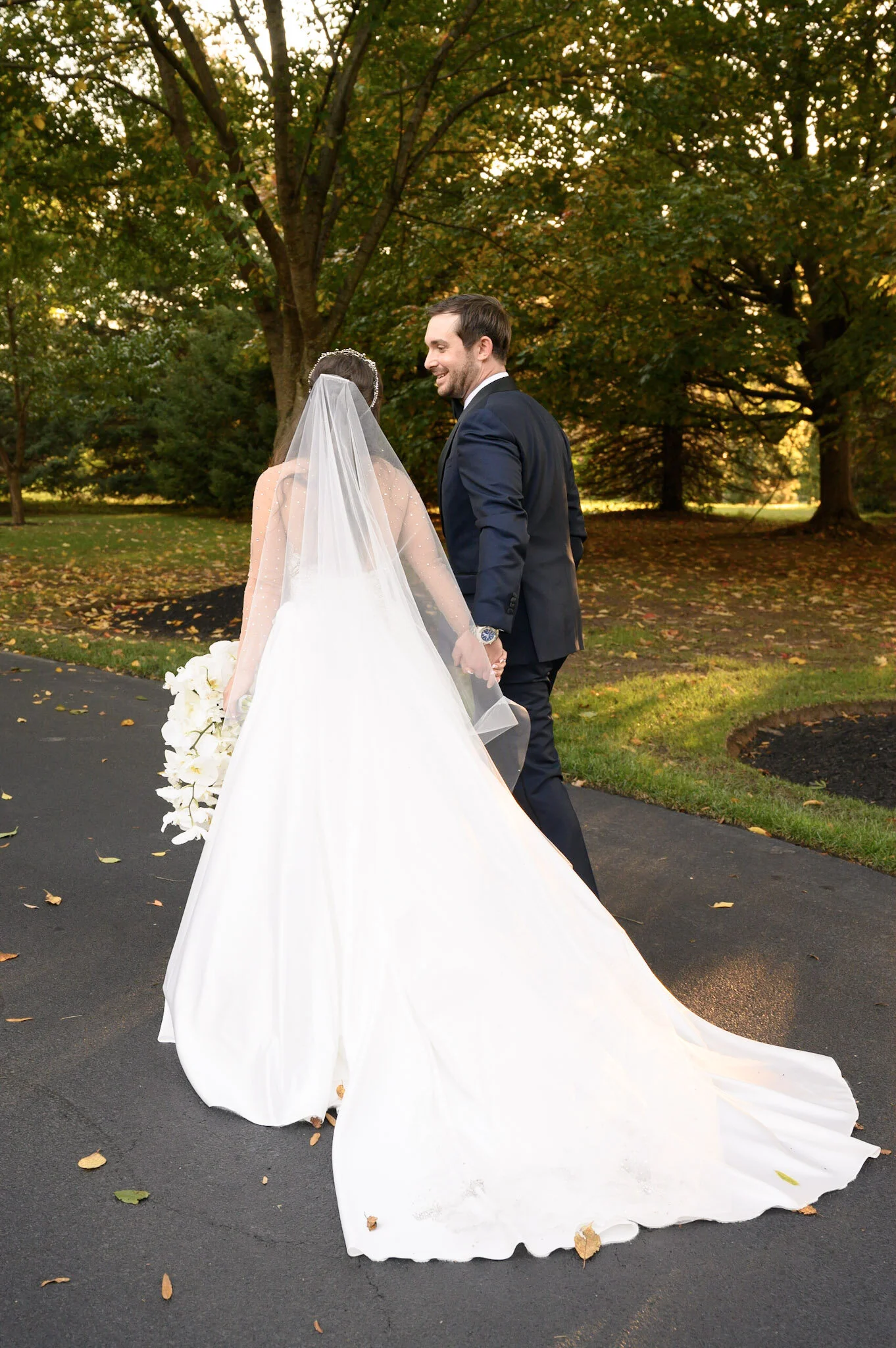 A bride and groom holding hands outdoors during their wedding, with the bride wearing a white gown with a long train and veil, and the groom in a black tuxedo, standing on a paved path surrounded by trees with green and yellow leaves.