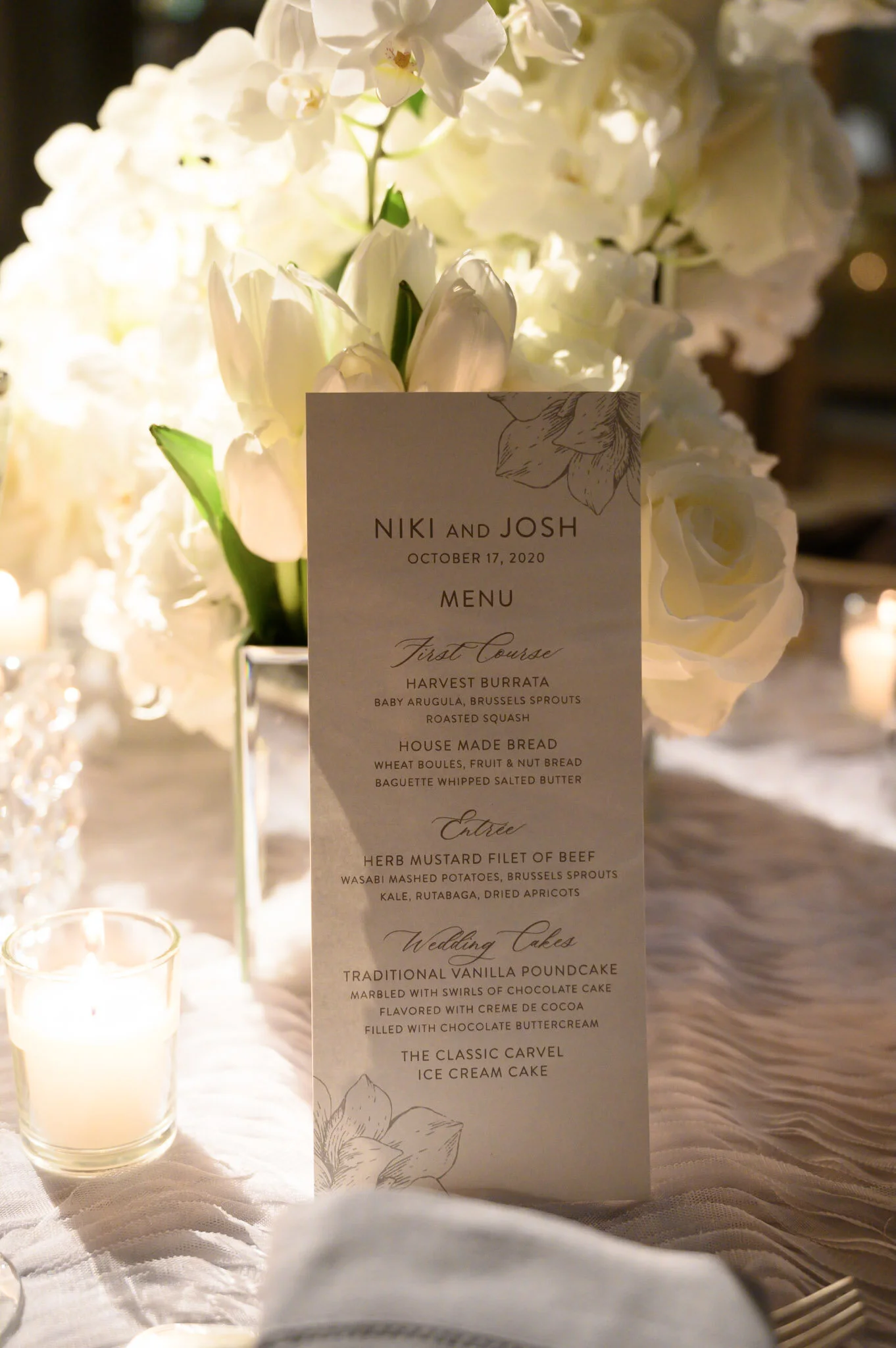 A wedding menu card with floral illustrations, placed in front of a background of white flowers. The menu includes first course, entree, and wedding cake options.