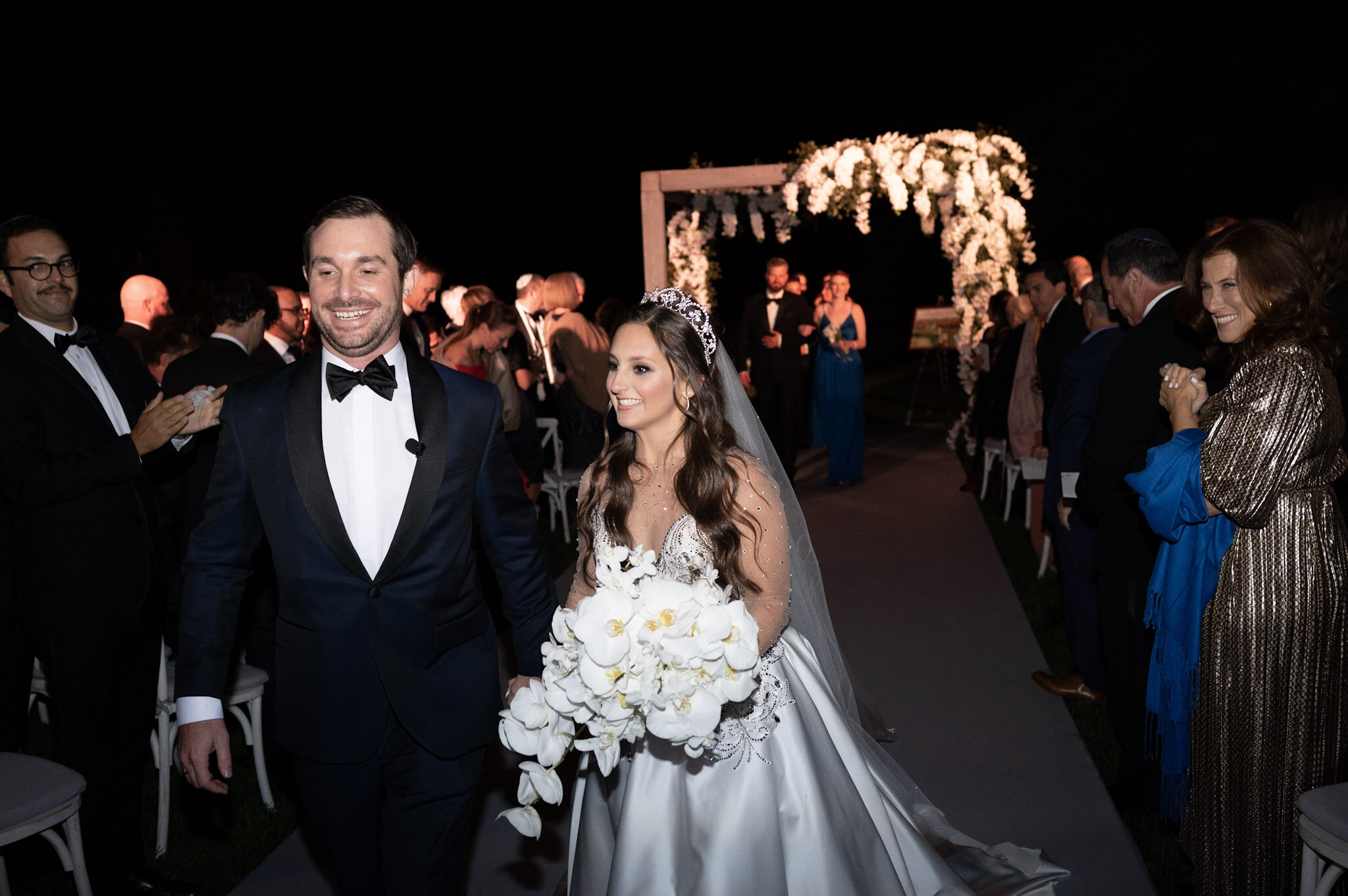 A bride and groom smiling as they walk down the aisle at a nighttime wedding reception, surrounded by guests under string lights and floral decorations.