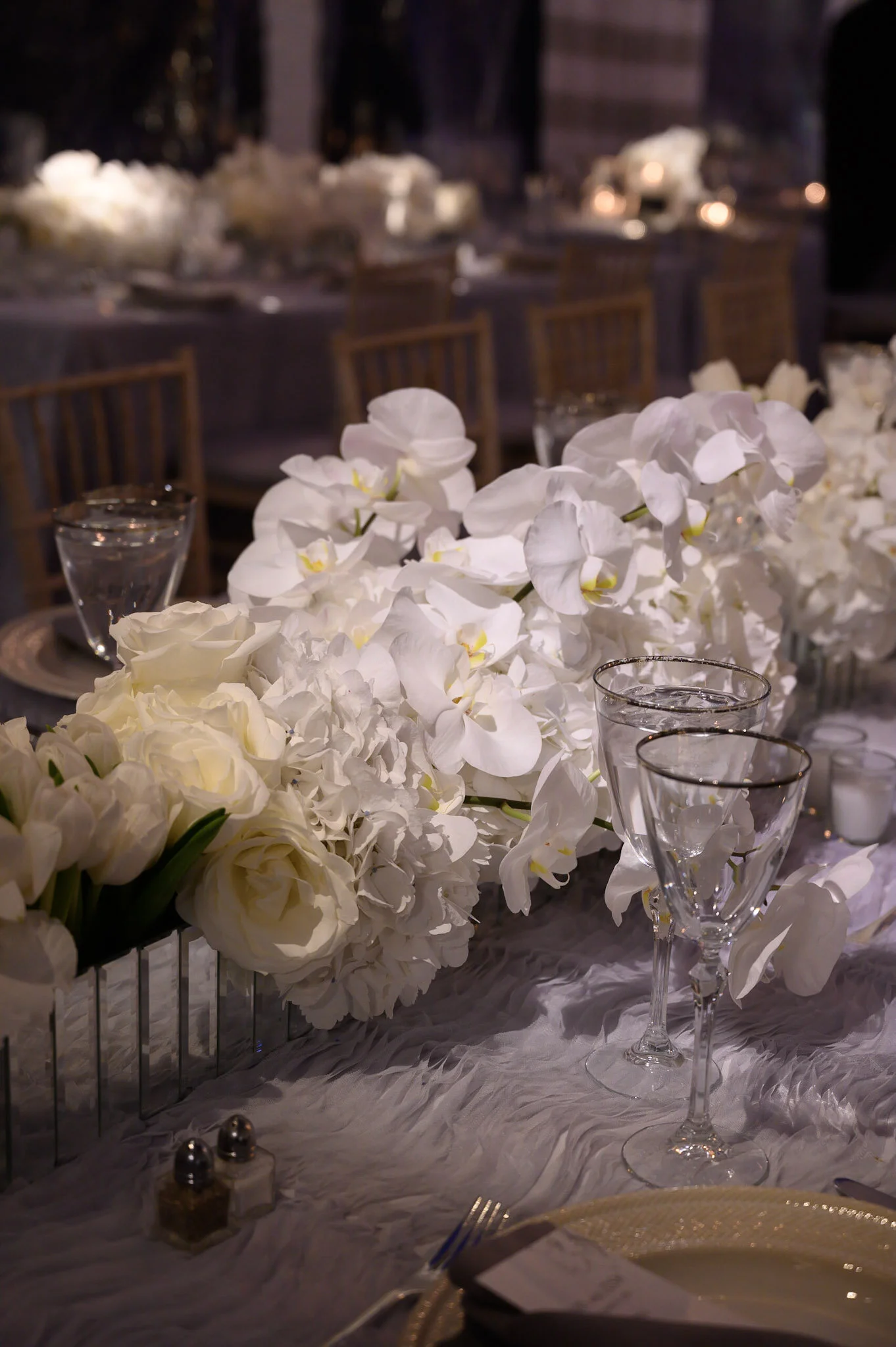 A close-up view of a table setting at a formal event with white flowers, clear glassware, and a golden plate, in a decorated dining area.