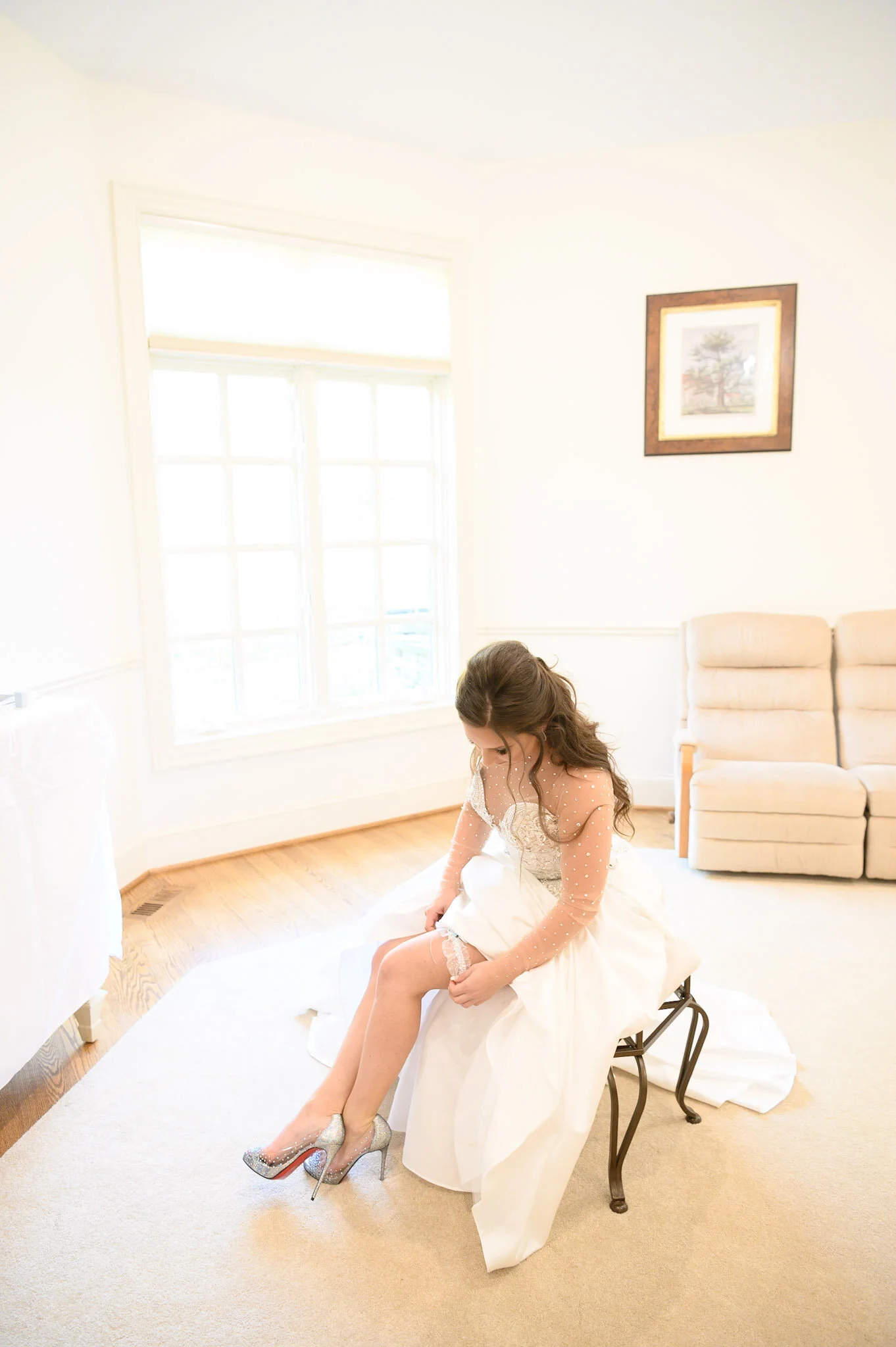A bride sitting on a chair while putting on her glittery high-heeled shoes in a bright living room with large windows and a framed painting on the wall.