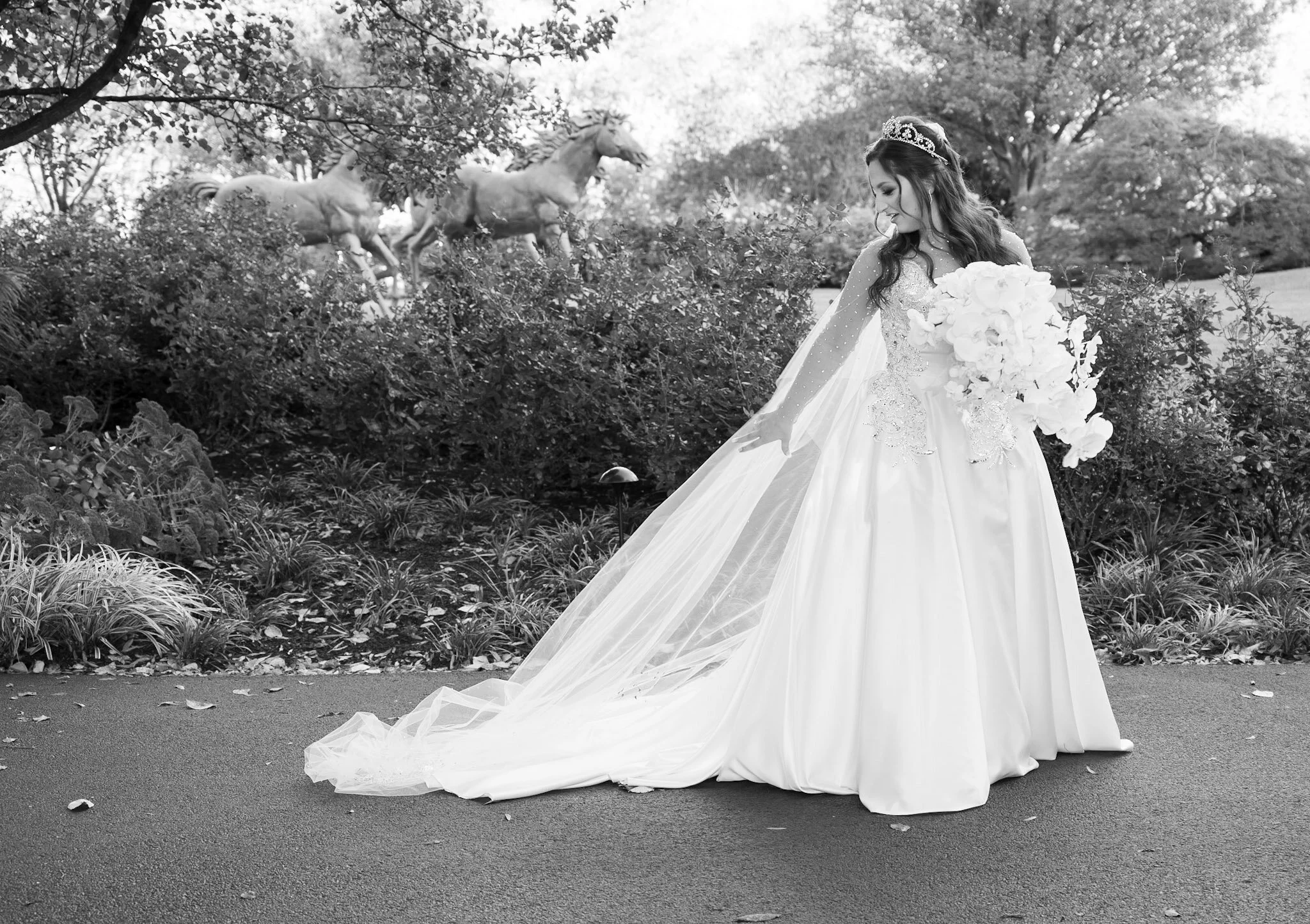 A black and white photo of a woman in a wedding dress with a long train, holding a bouquet of flowers, and wearing a tiara. She is outdoors, with trees and bushes in the background and two horses in the distance.