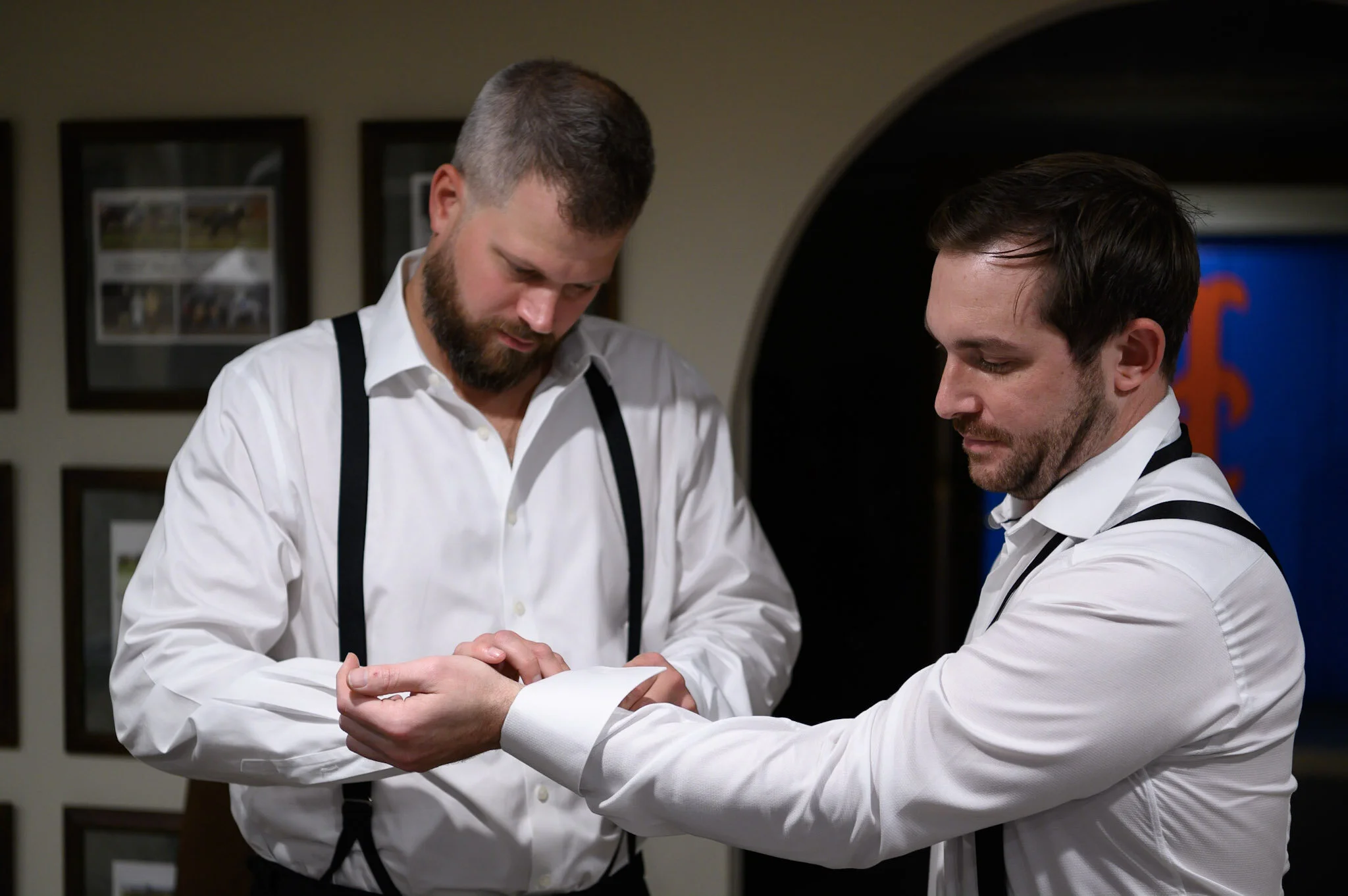 Two men in white shirts and black suspenders are looking at each other's hands. They are standing indoors, with framed pictures on the wall behind them.