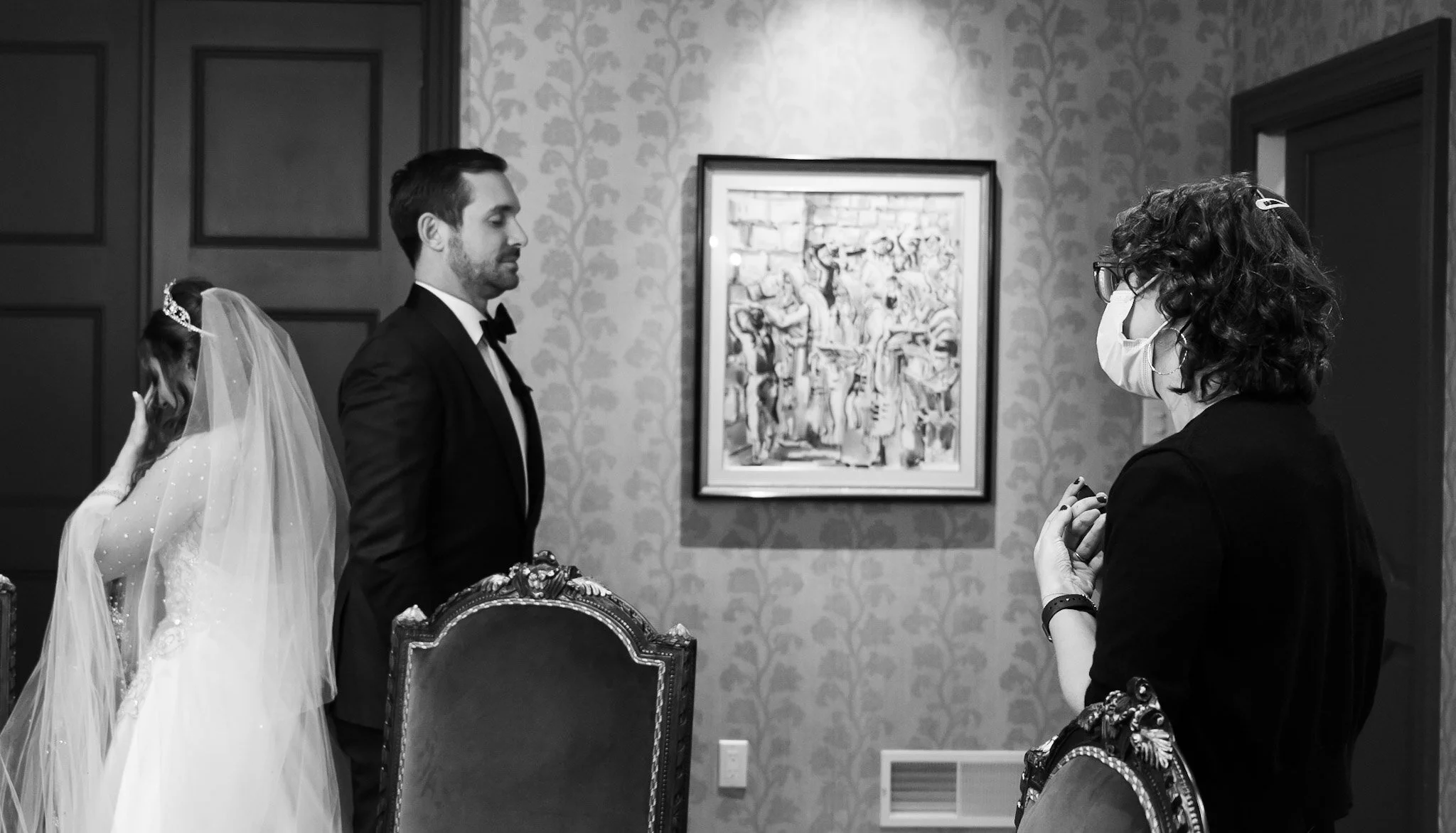 Black and white photo of a wedding scene with a bride in a wedding dress with veil, groom in a tuxedo, and two women, one wearing glasses and a face mask, inside an art gallery or museum with patterned wallpaper and artwork on the wall.