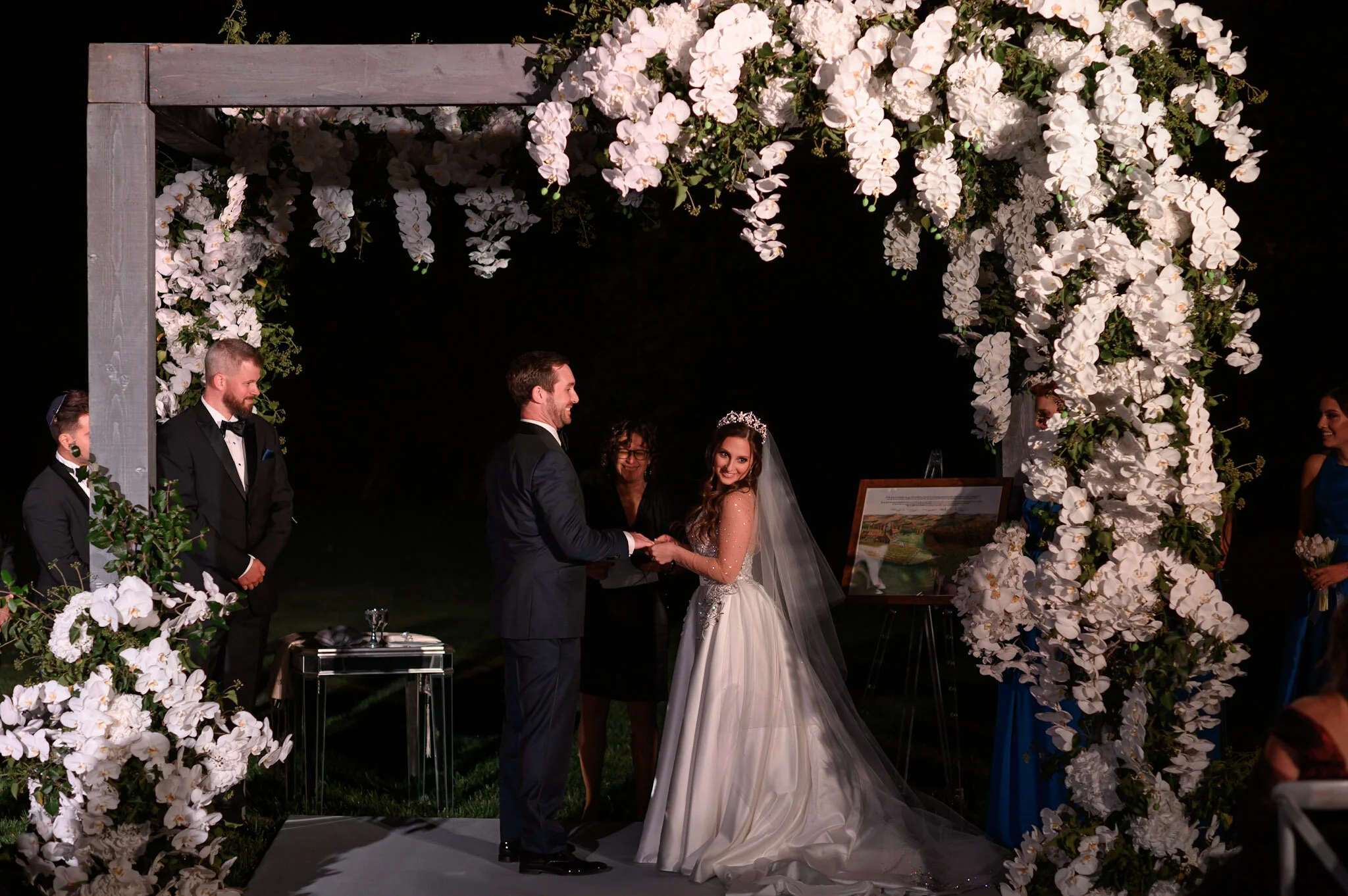 A wedding ceremony taking place under a floral arch with the bride and groom holding hands and smiling at each other. Bridesmaids and groomsmen are standing on either side, with the bride in a white dress and veil, and the groom in a dark suit. The a