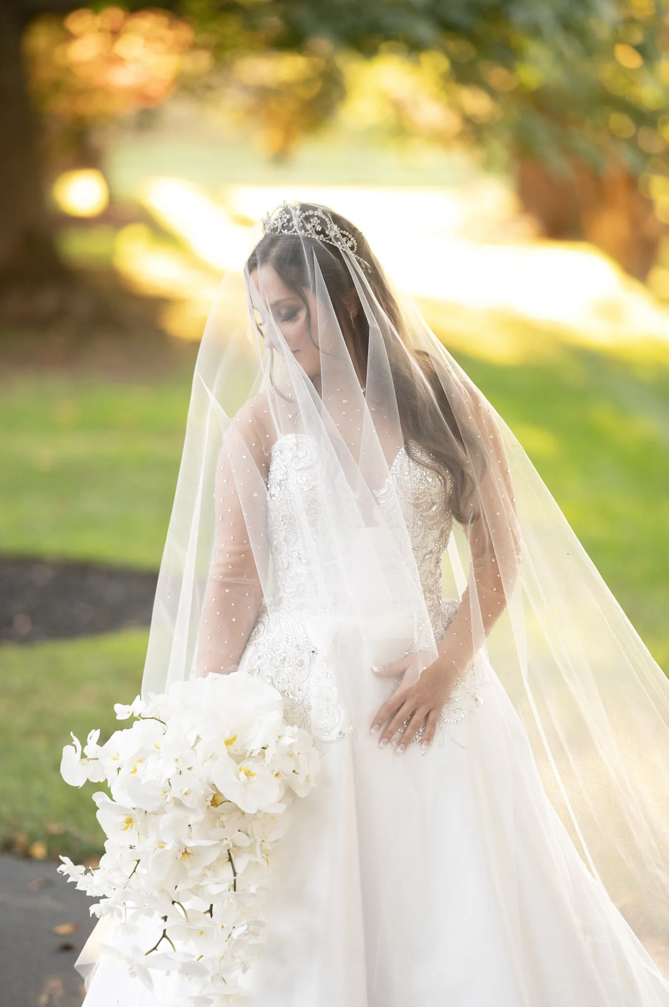 Bride in a white wedding gown and veil holding a large bouquet of white orchids, outdoors with a background of trees and sunlight.