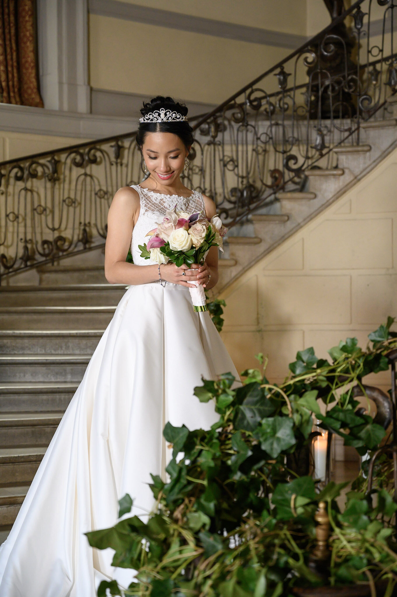 A bride wearing a white wedding gown and tiara, holding a bouquet of roses, standing on a staircase inside a building with ornate iron railings and decorative surroundings.