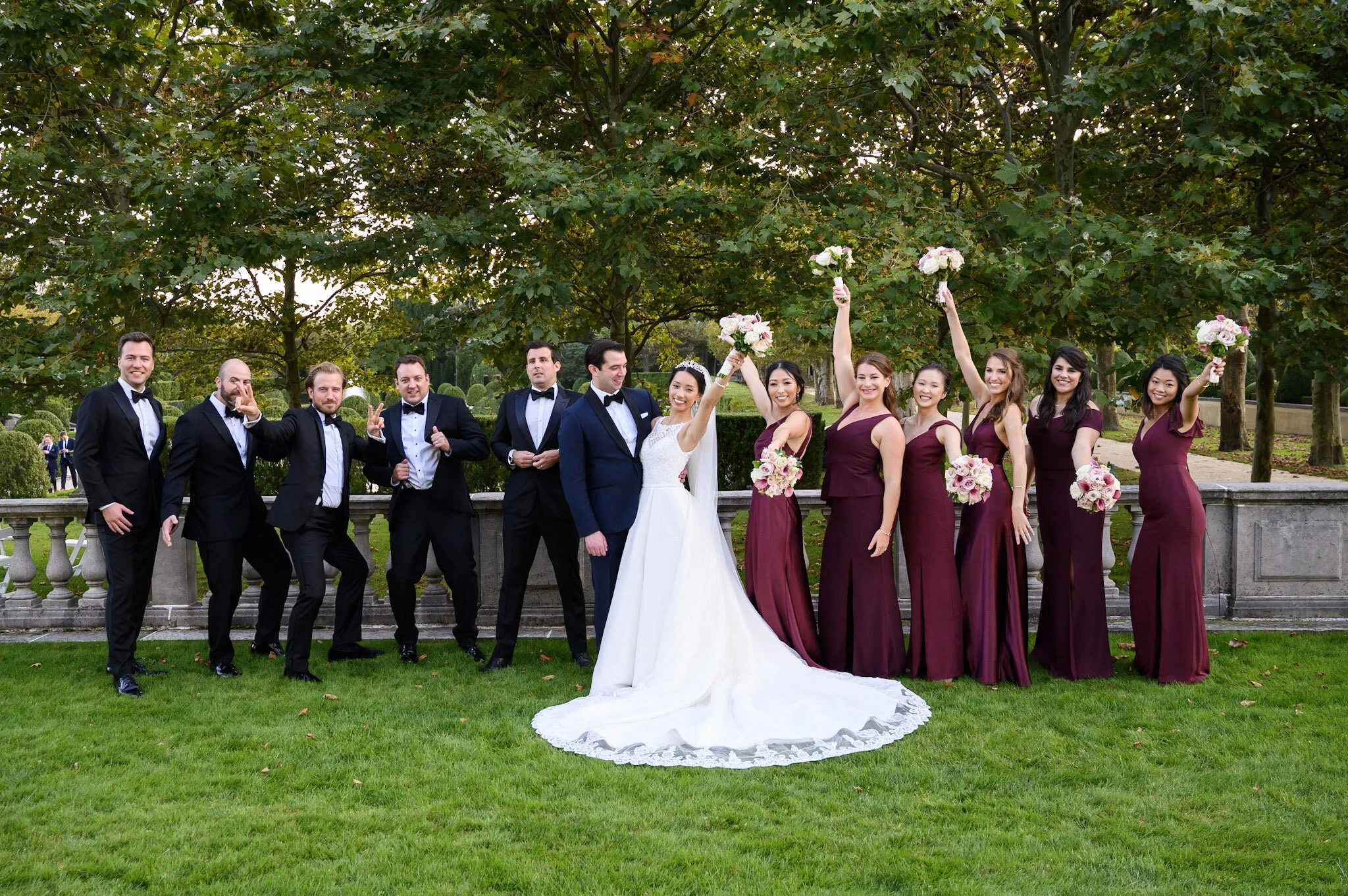 Group of people in wedding attire, smiling and posing outdoors under trees, with some women holding bouquets of pink and white flowers, during daytime.