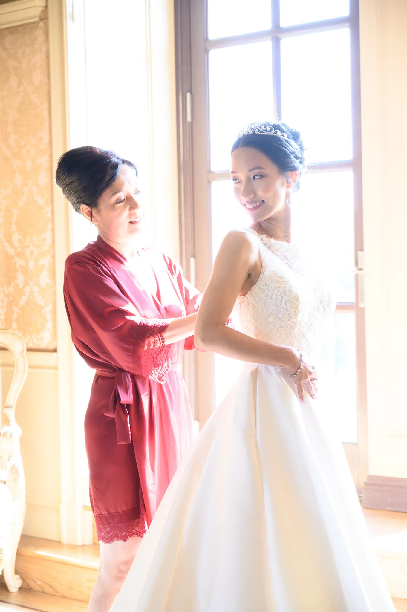 A bride in a white wedding dress with lace details, wearing a tiara, smiling and looking to her right while an older woman, possibly her mother, helps her with her dress. The scene is bright with sunlight streaming through a large window in the backg