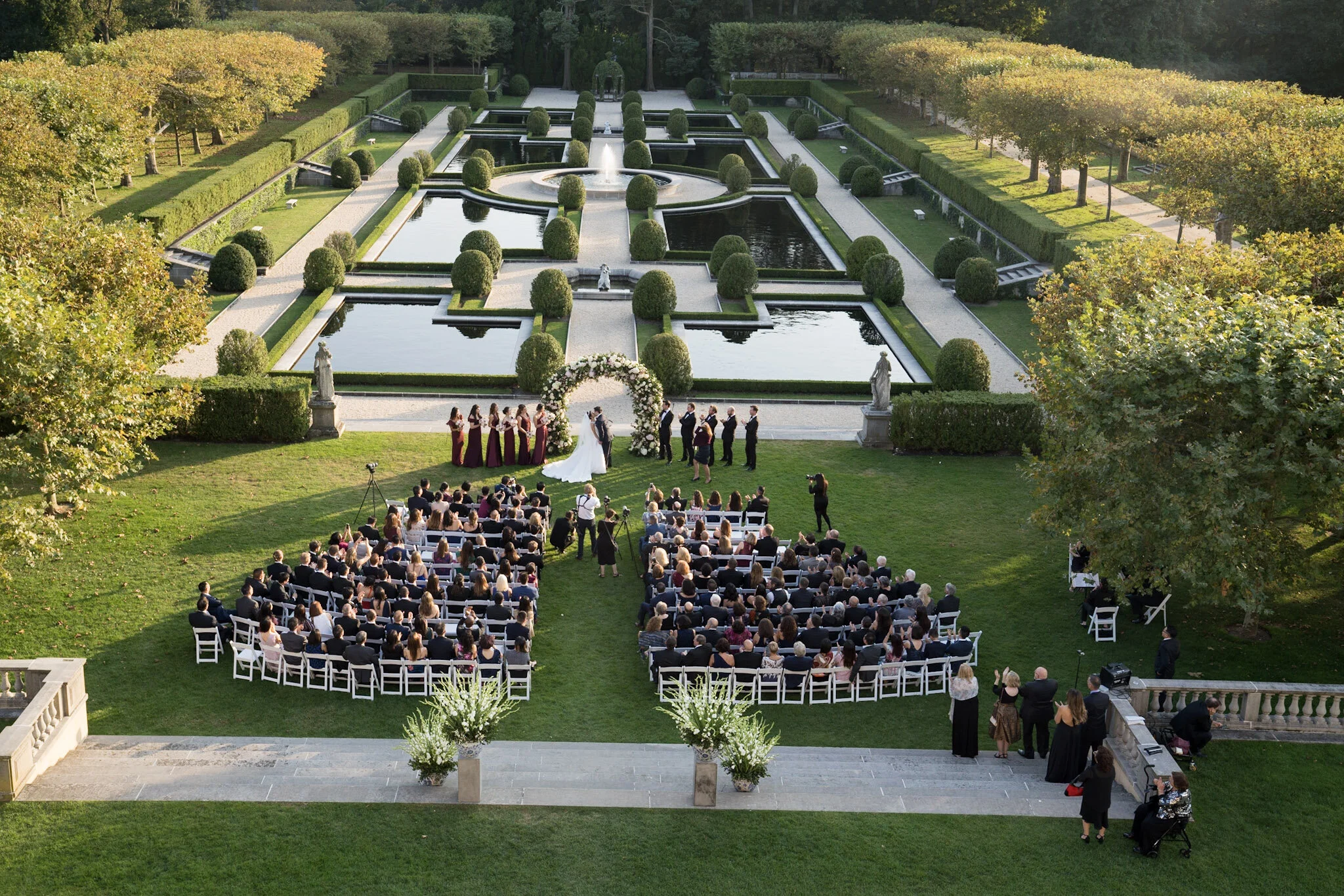 An outdoor wedding ceremony taking place on a lush green lawn with an elaborate geometric water fountain garden backdrop. Guests are seated on white chairs, facing a bride and groom under an floral arch, with bridesmaids and groomsmen standing nearby