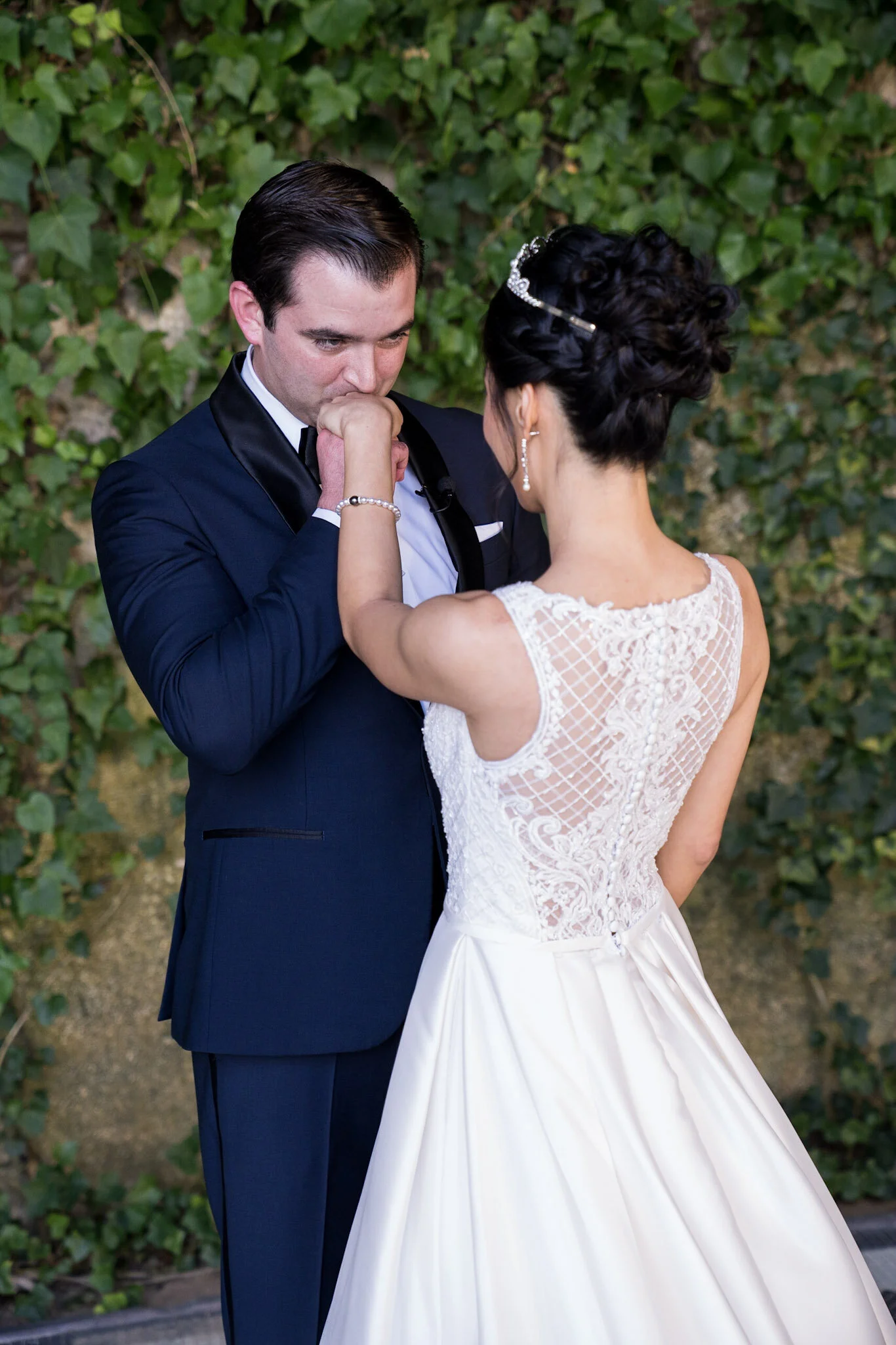 A bride and groom share an intimate moment outdoors with green leafy background. The groom is dressed in a dark blue tuxedo and the bride in a white lace wedding gown with her hair styled in curls and a tiara.
