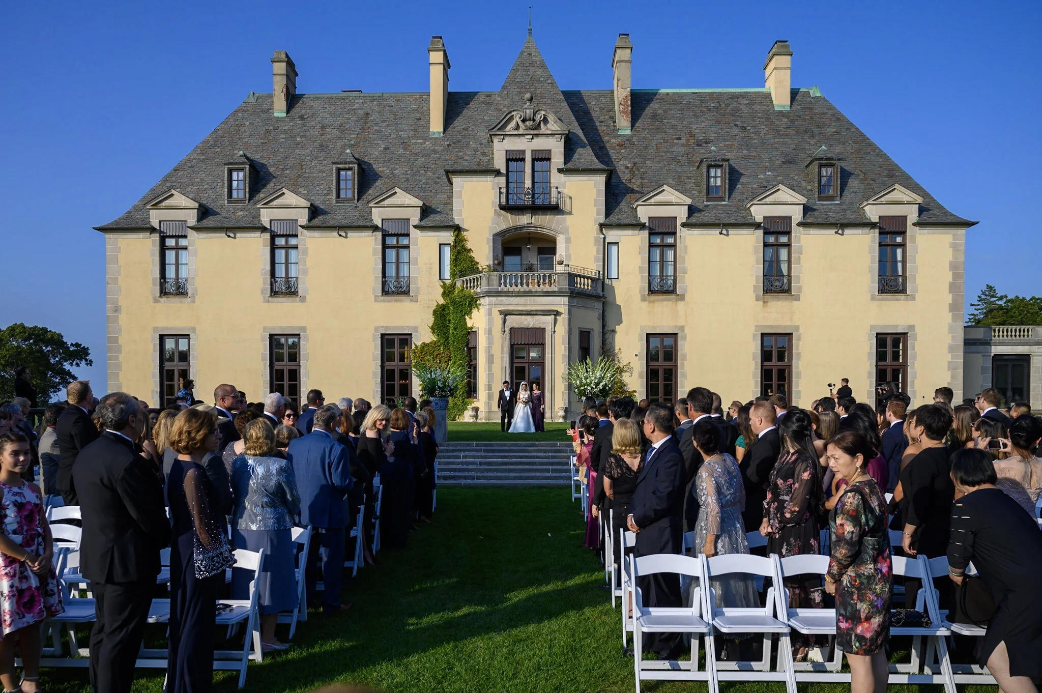 A large group of people gathered outdoors for a wedding ceremony in front of a historic mansion or castle with a gray stone roof, tall chimneys, and decorative architectural features. The bride and groom stand at the top of stairs under an archway, w