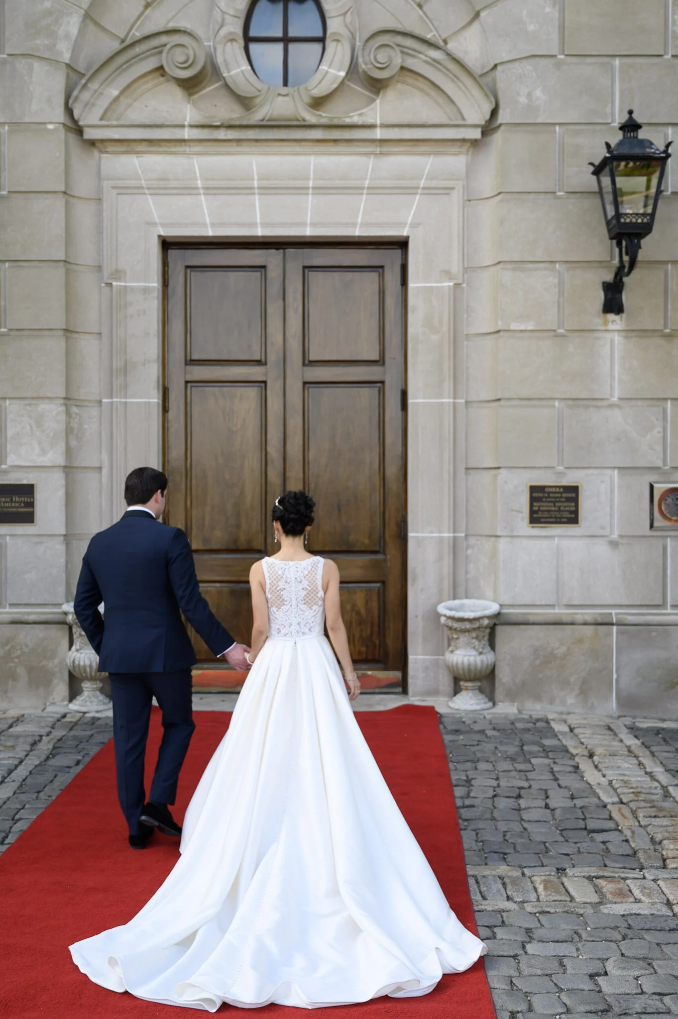 A bride and groom holding hands walking down a red carpet outside a stone building with a large wooden door and a decorative window above it.