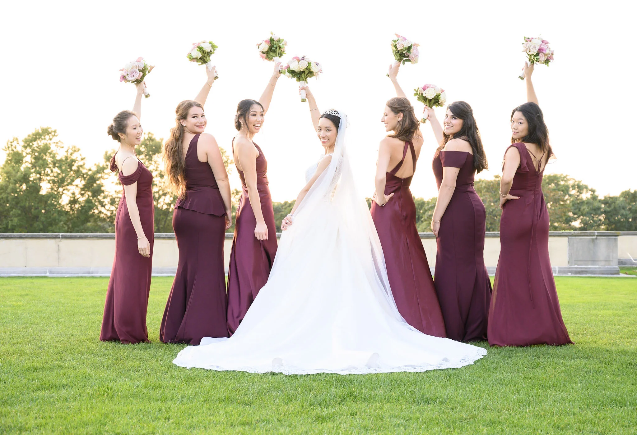 A bride and six bridesmaids standing on a grassy field, all holding bouquets of pink and white flowers and smiling. The bride wears a white dress with a veil and a tiara, while the bridesmaids wear matching purple dresses. They are celebrating outdoo