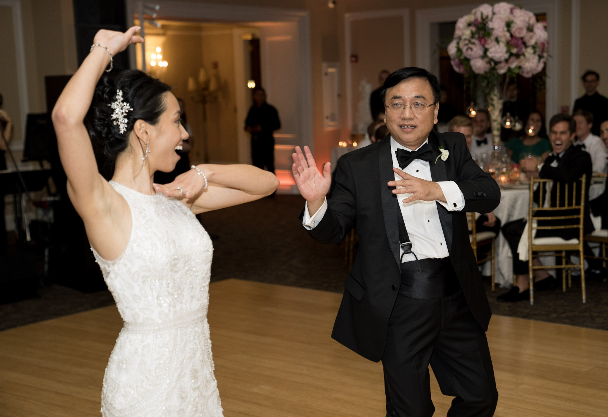 A bride and an older man, likely her father or a close relative, dancing at a wedding reception. The bride is wearing a white lace wedding gown with a floral hair accessory, and the man is in a black tuxedo with a bow tie.