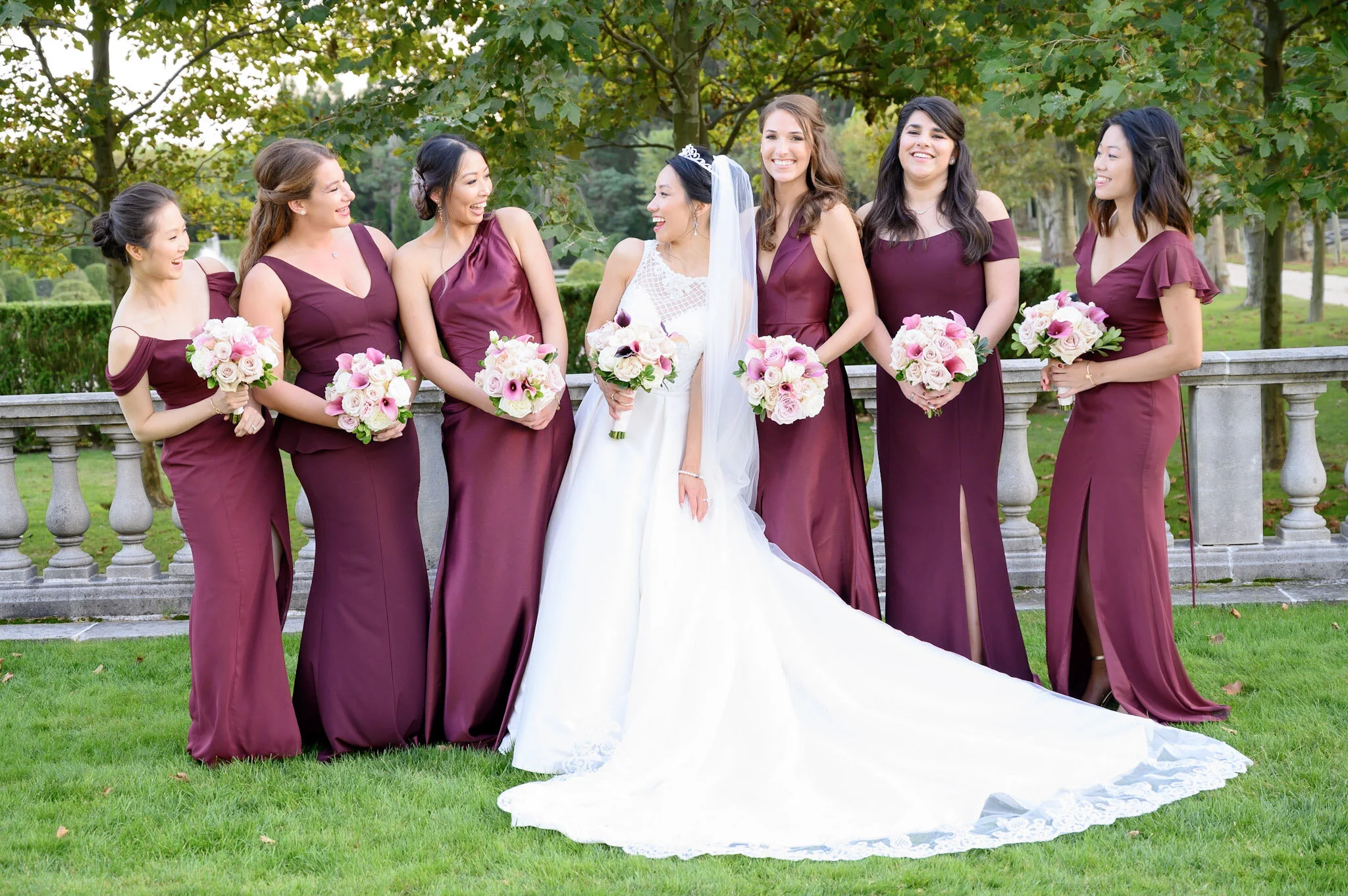 Group of seven women dressed in maroon dresses, one in a white wedding gown with a veil and tiara, standing outdoors on a grassy area with trees, holding bouquets of pink and white flowers, smiling and looking at each other.