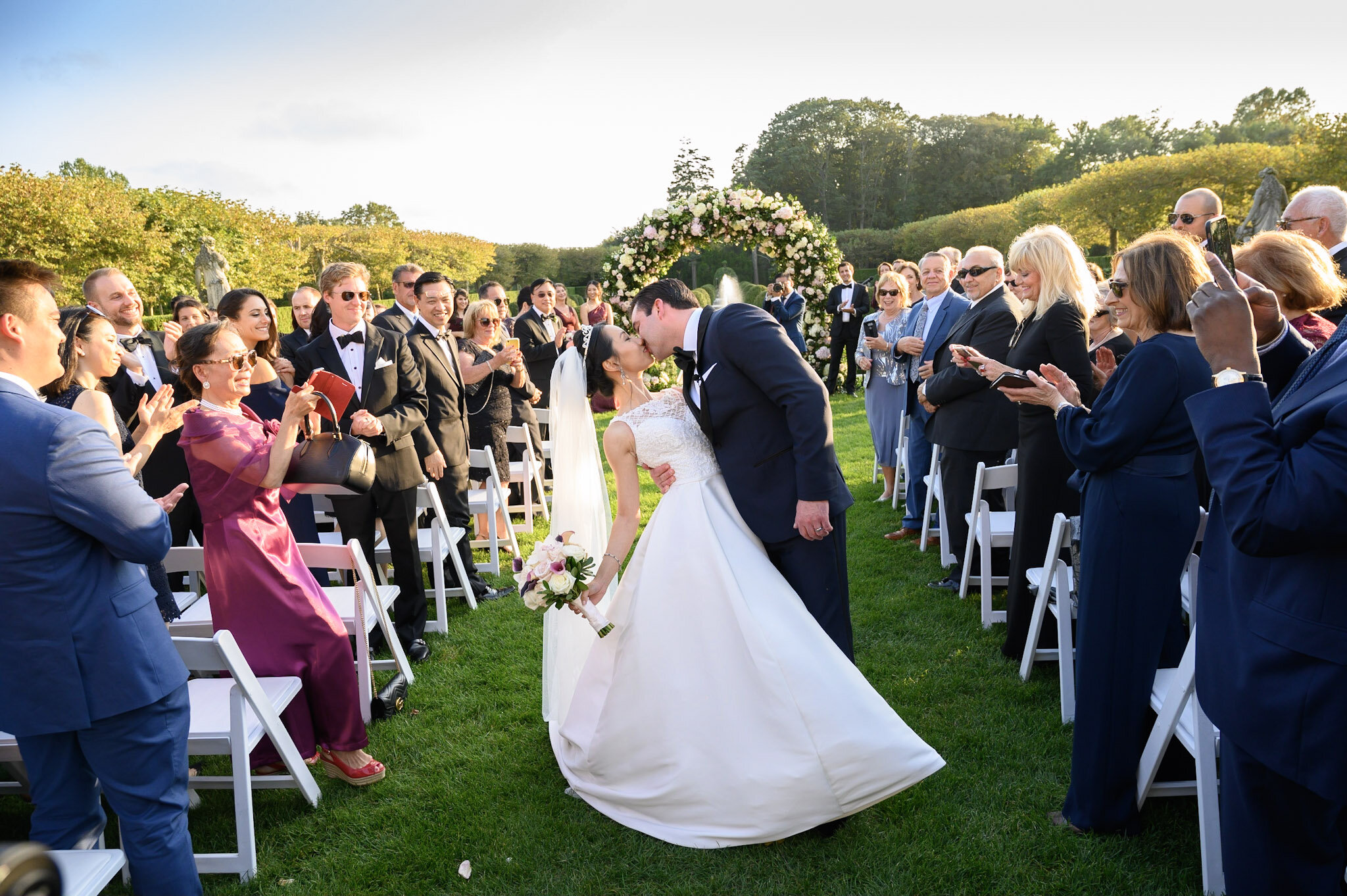 A wedding ceremony outdoors with a bride and groom kissing under an arch of flowers, surrounded by seated guests on a sunny day.