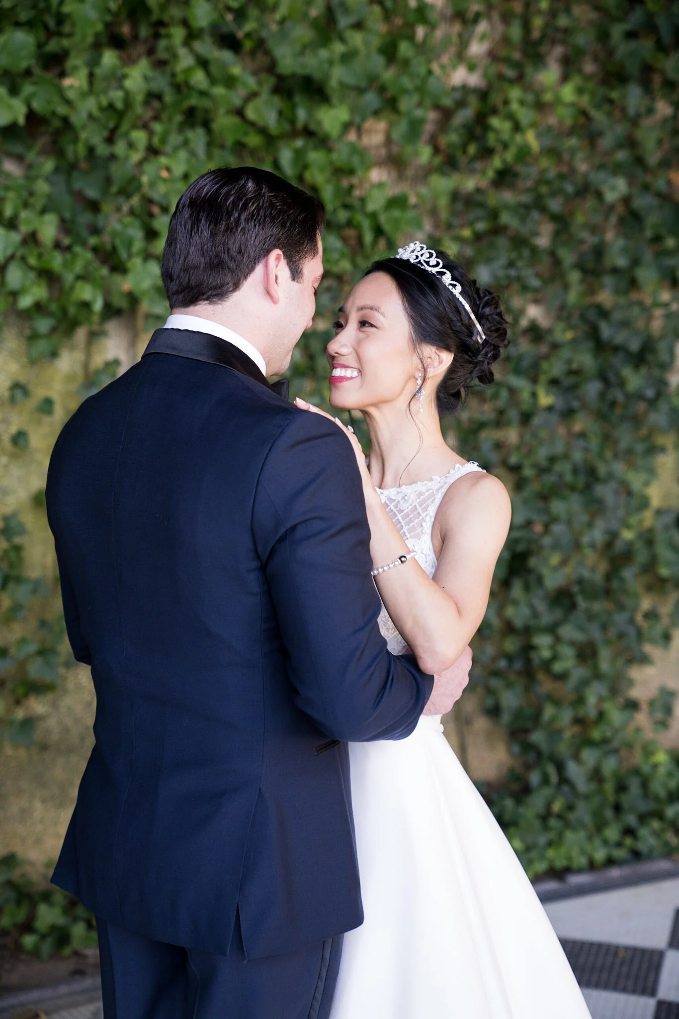 A bride and groom are embracing outdoors against a green leafy background. The bride is wearing a white wedding dress and a tiara, and the groom is in a dark suit. Both are smiling and looking into each other's eyes.