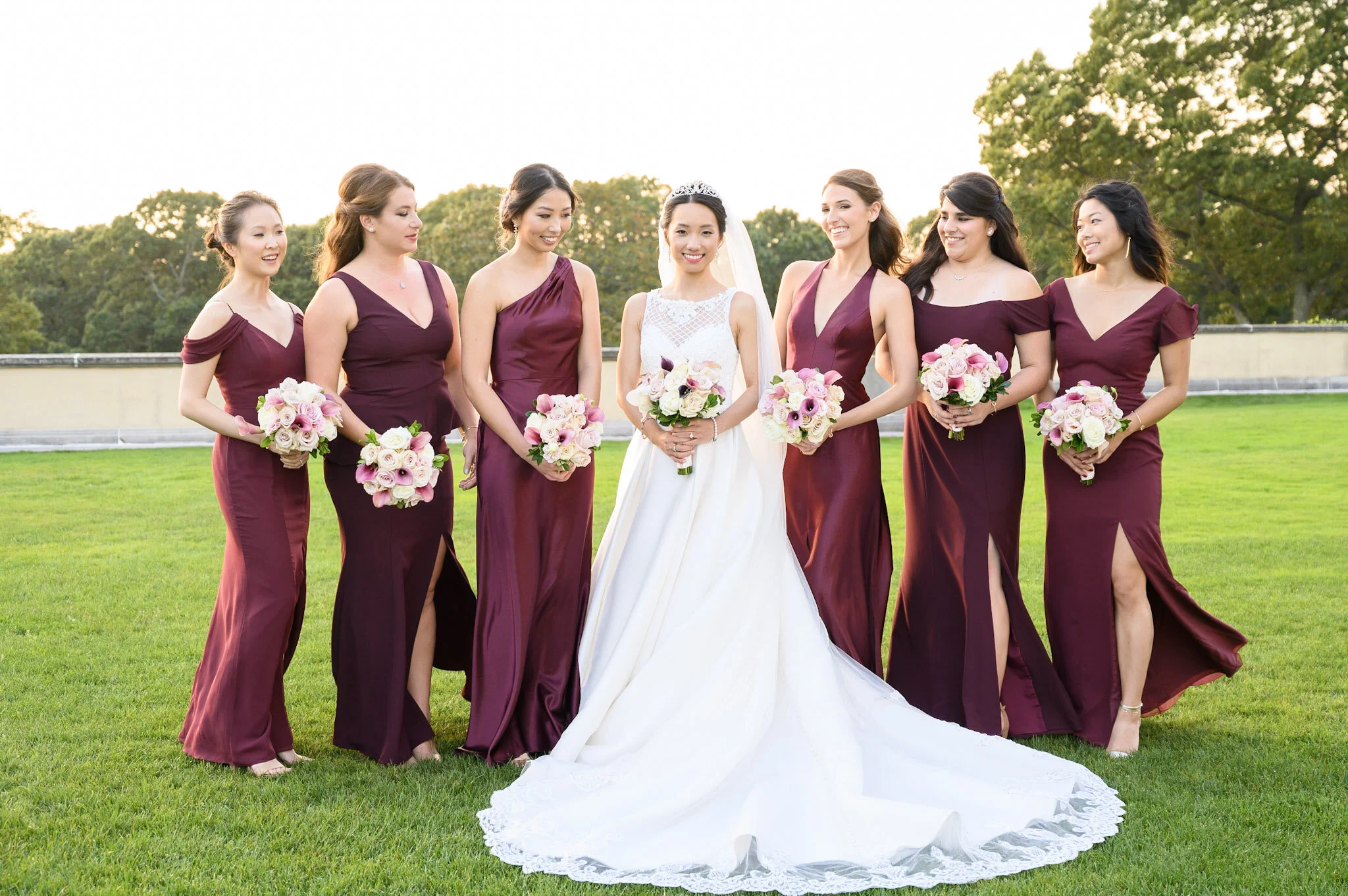 A bride in a white wedding dress and tiara standing with six bridesmaids in matching burgundy dresses on a grassy field with trees in the background.