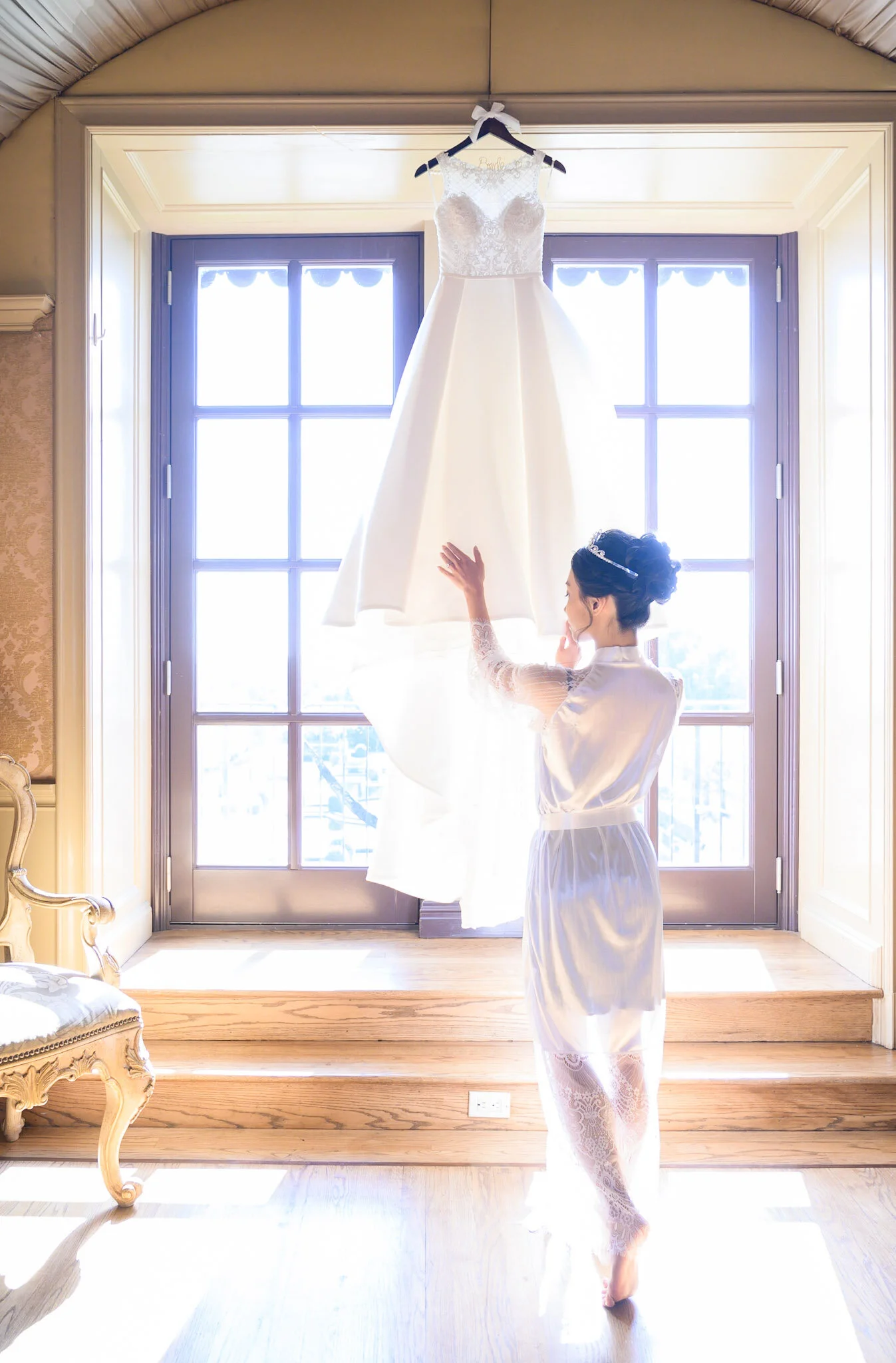 A bride in a satin robe and lace stockings stands inside near a large window, reaching up to touch her wedding dress that hangs on a hanger against the window.