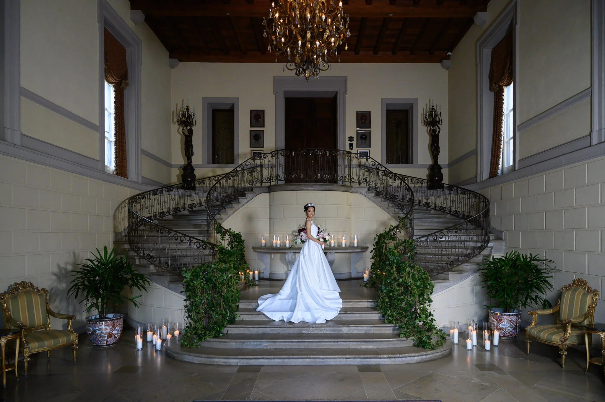 A bride in a white wedding gown and tiara standing on a grand staircase decorated with candles and green foliage, inside an elegant mansion with chandeliers and ornate furniture.