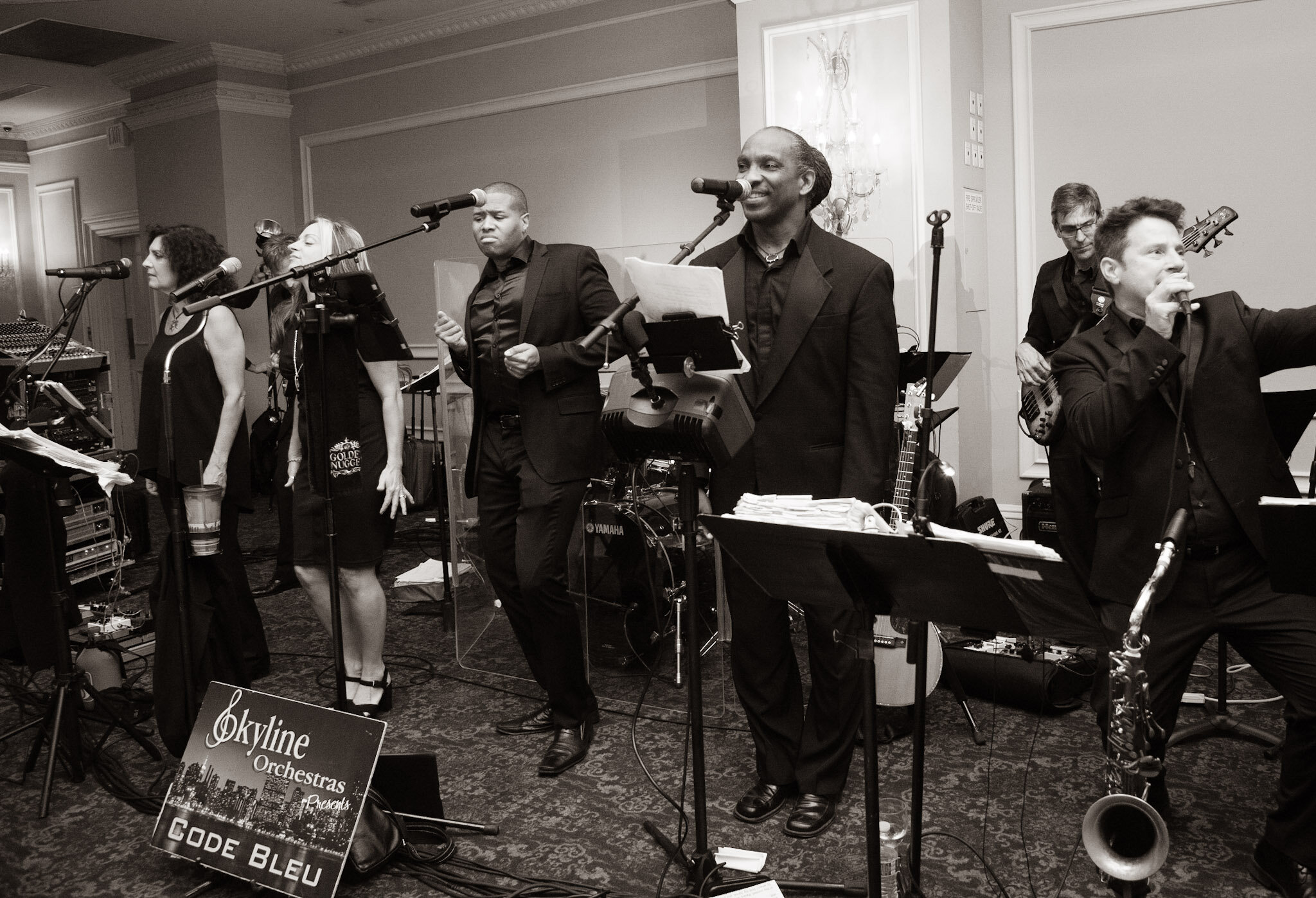 A group of musicians performing at an indoor event, with several people singing into microphones and playing instruments, including a bass guitar and a keyboard, in a formal setting with ornate decorations on the walls.