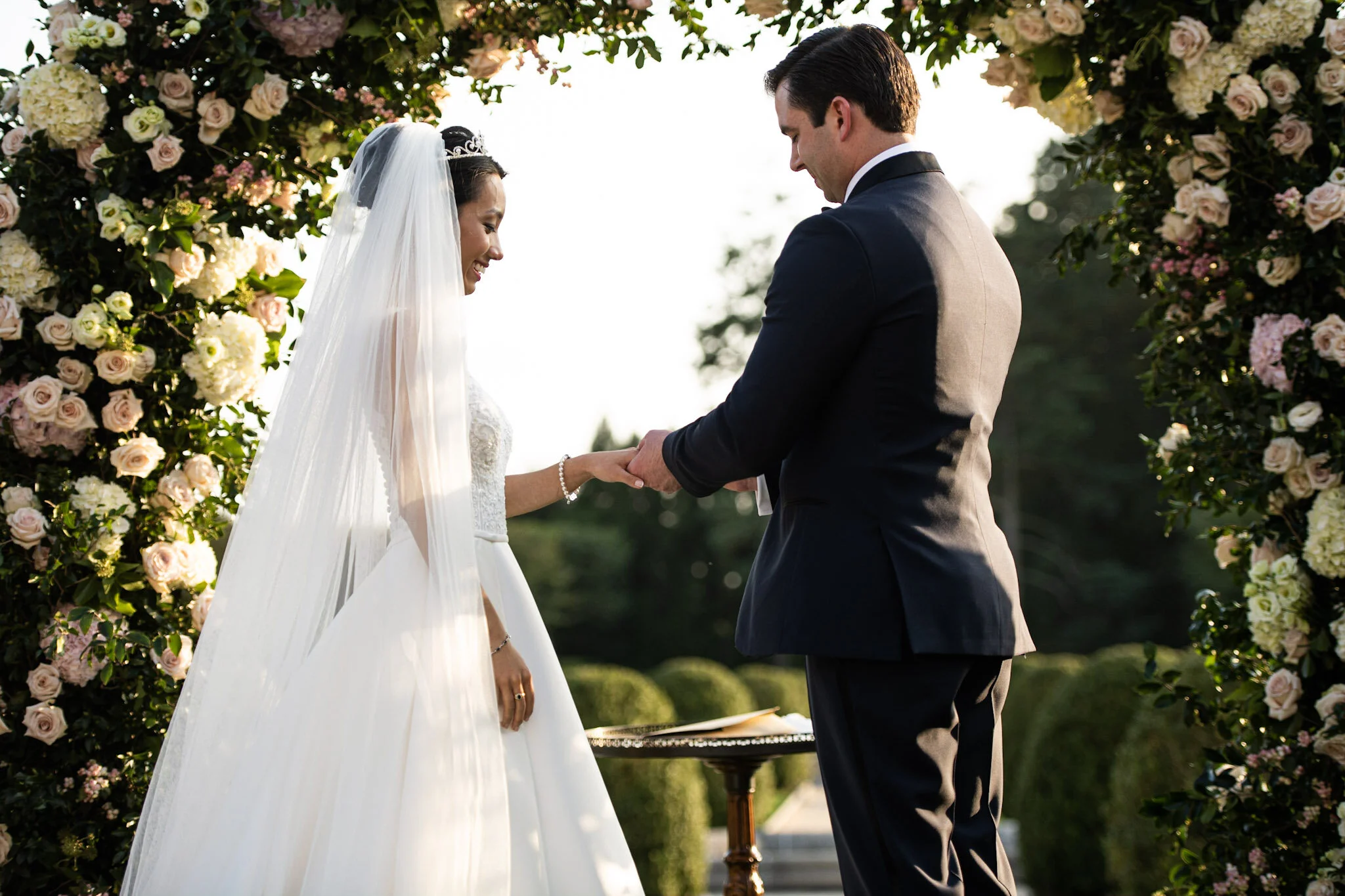 A wedding ceremony with a bride and groom holding hands under a floral arch, smiling, with greenery in the background during sunset.