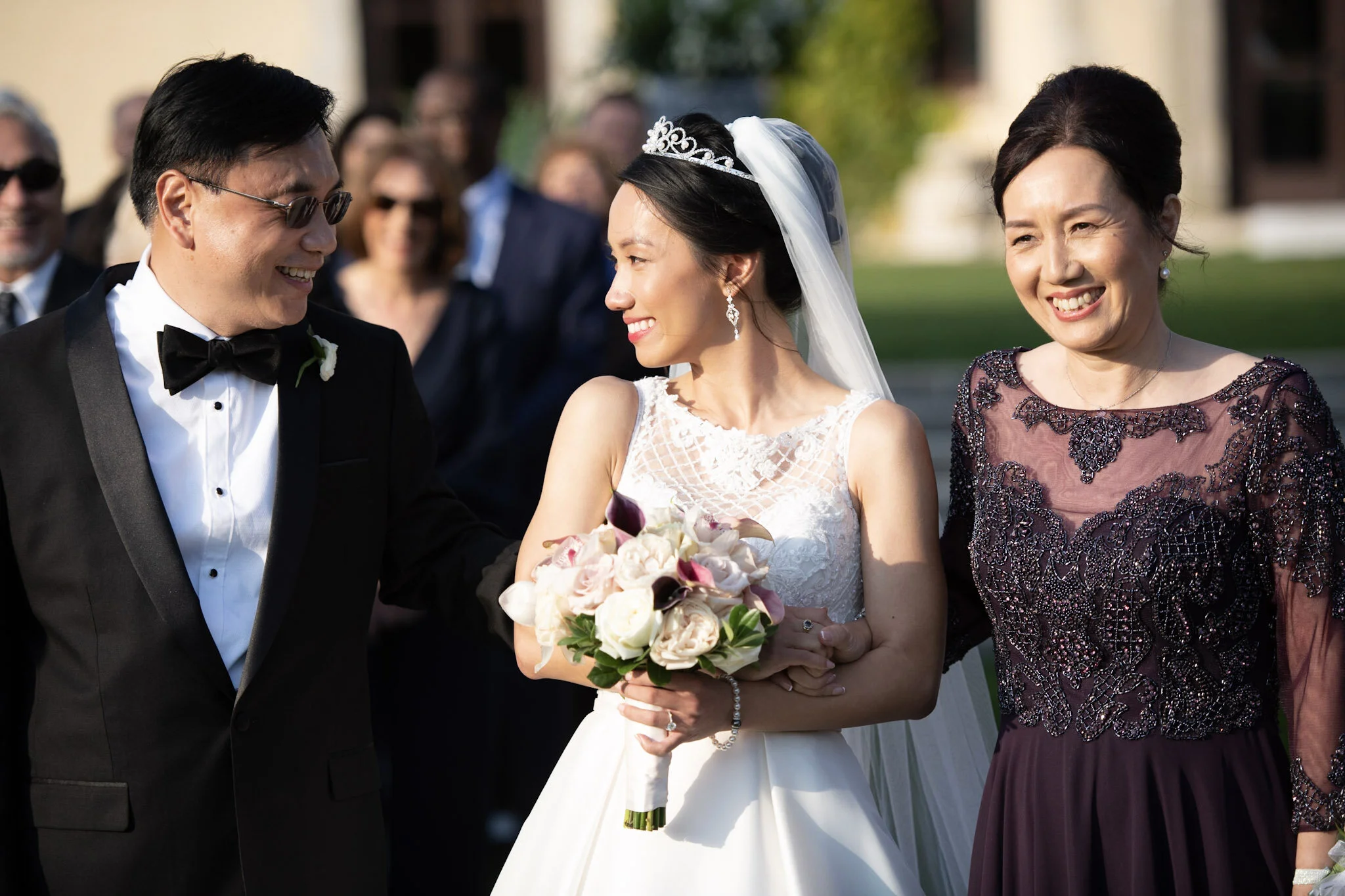 Bride in white wedding dress holding bouquet and smiling at groom dressed in black tuxedo with black bow tie, walking outdoors with a woman in a dark purple dress and pearl earrings.
