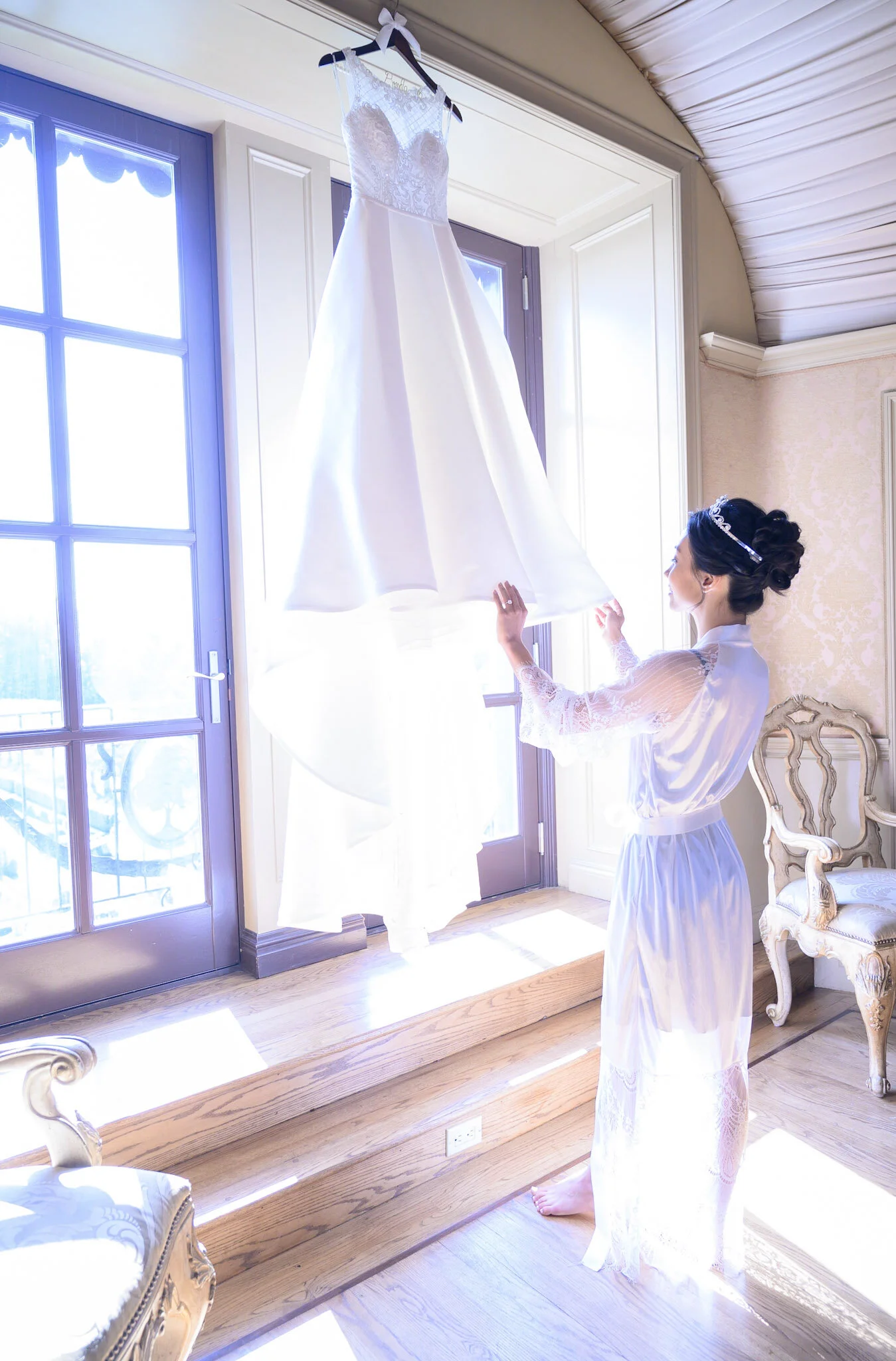 A woman in a white robe with lace details stands indoors, holding a wedding dress hanging near a large window with sunlight streaming in.