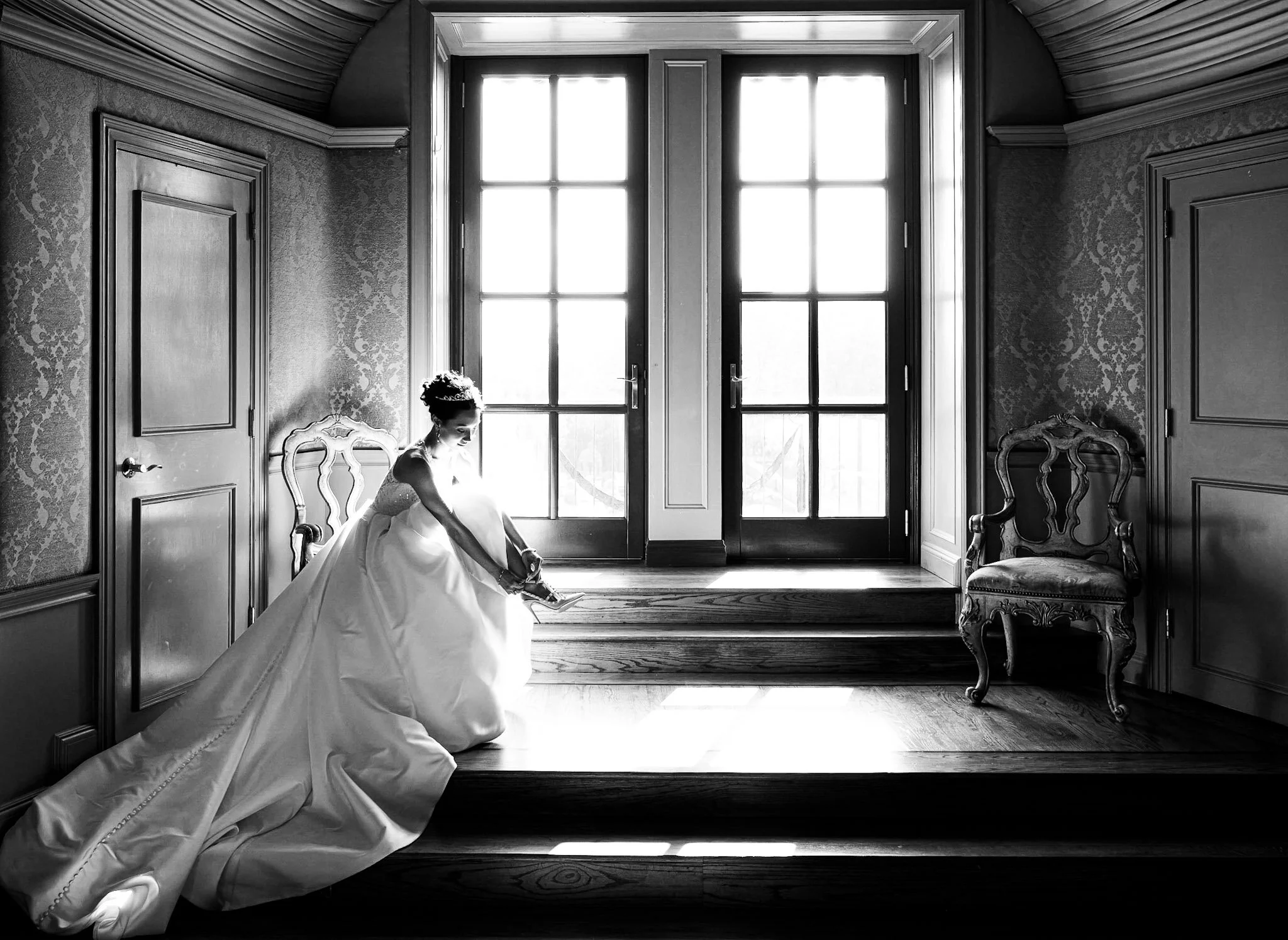 A bride sitting on a chair by large windows, putting on her high heel shoe. The room has elegant wallpaper, closed doors, and vintage furniture.