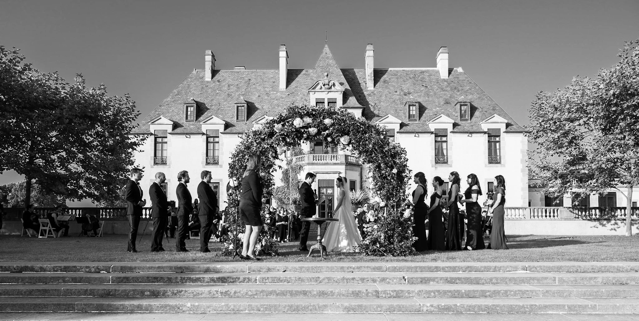 A black-and-white photo of a wedding ceremony taking place outdoors in front of a large, elegant house. The bride and groom stand facing each other beneath a floral arch with bridesmaids and groomsmen lined up on either side. Guests sit in the backgr