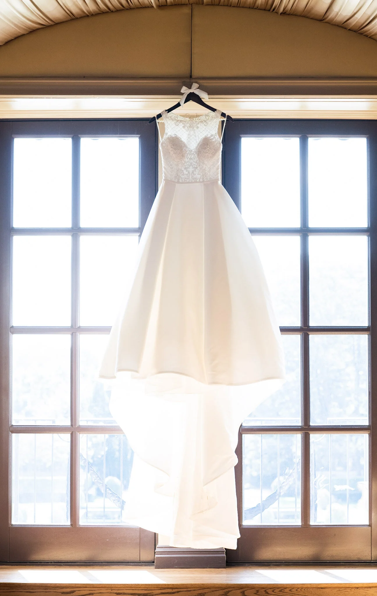 White wedding dress hanging on a hanger in front of a glass door, backlit by sunlight.