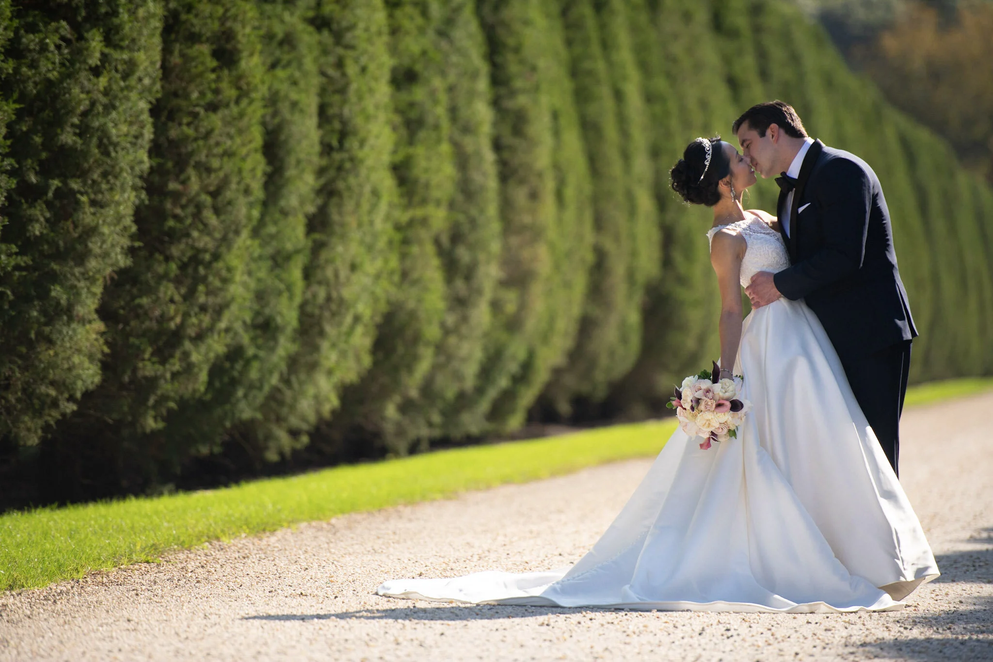 A bride and groom sharing a kiss outdoors in a garden setting, with the bride holding a bouquet of flowers. The bride is wearing a white wedding gown and a tiara, and the groom is in a black tuxedo with a bow tie. The background features green hedge 