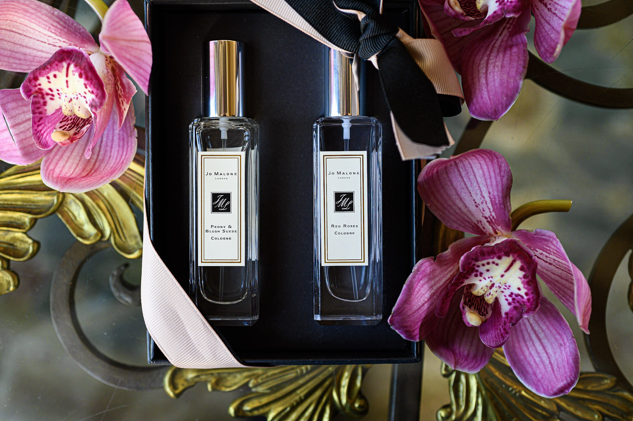 Two bottles of Jo Malone cologne, labeled 'Peony & Blush Suede' and 'Red Roses', are placed inside a black box with a pink ribbon. The box is surrounded by pink orchids and golden decorative elements.