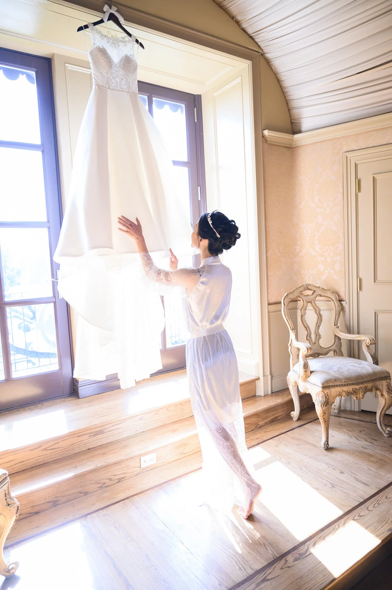 A woman in a lace robe touches a white wedding dress hanging in front of a window inside a room with wooden floors, cream-colored walls, and an antique chair.