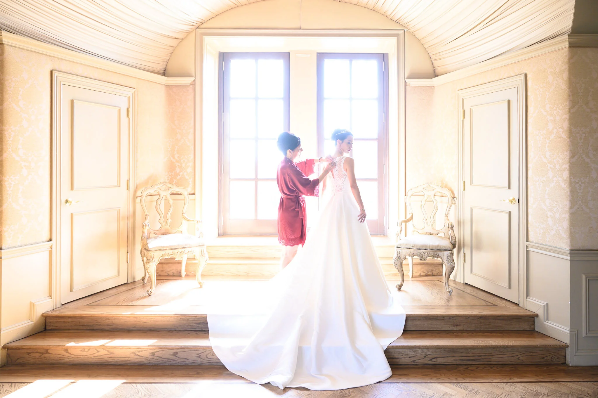 A bride in a white wedding gown and veil standing by a large window with sunlight streaming in, assisted by a woman in a red robe, in a decorated room with cream walls and ornate chairs.