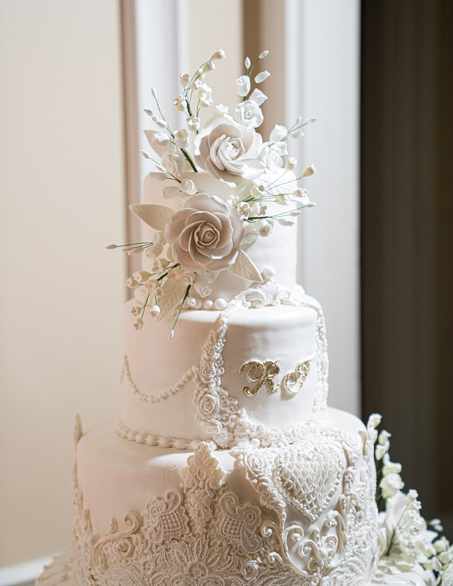 A white wedding cake decorated with intricate piping lace designs, white roses, and baby's breath flowers. The cake has gold initials "K" and "S" on the front.