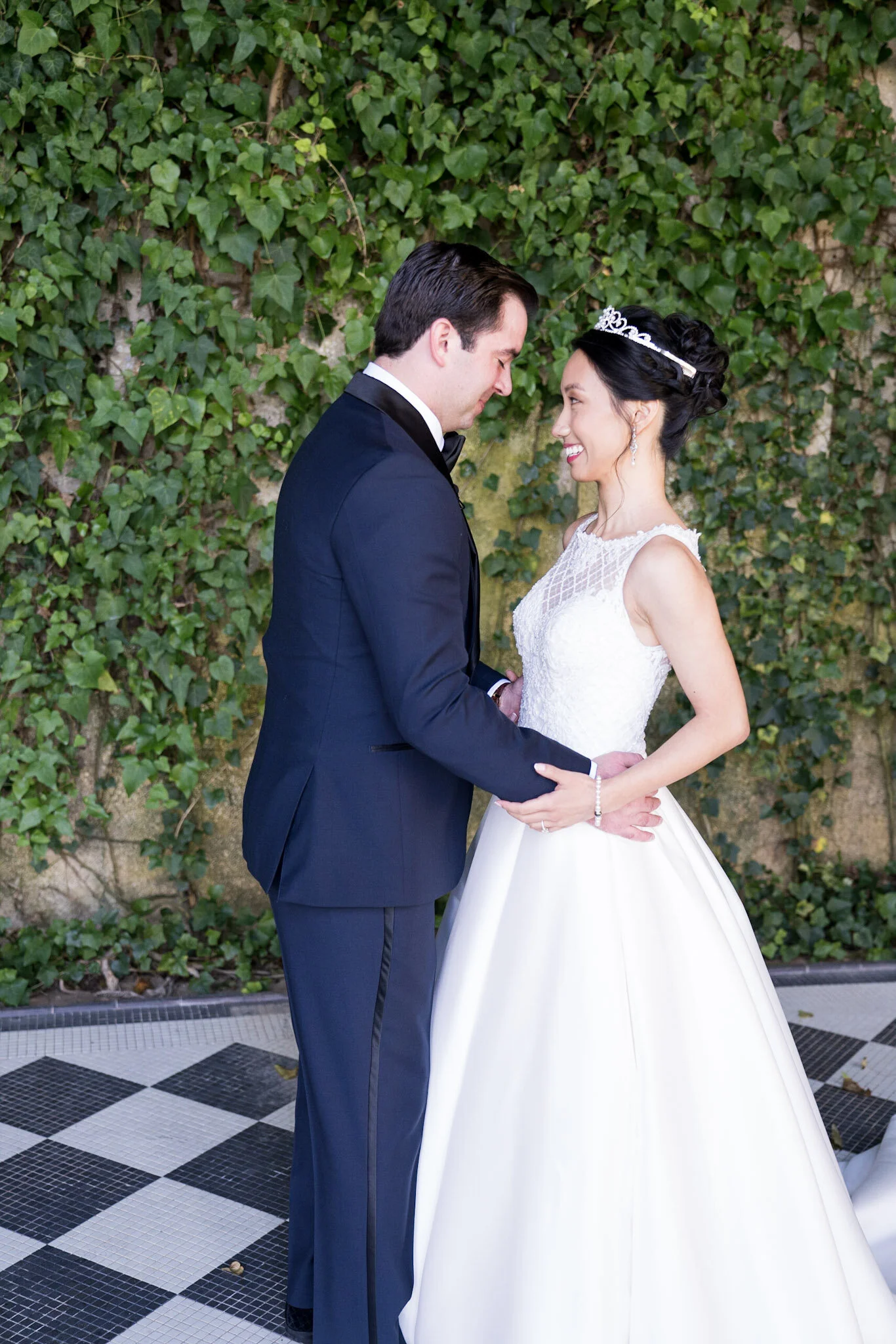 A bride and groom stand close together, smiling and touching foreheads, against a green leafy wall. The groom wears a dark tuxedo, and the bride wears a white wedding gown and tiara.