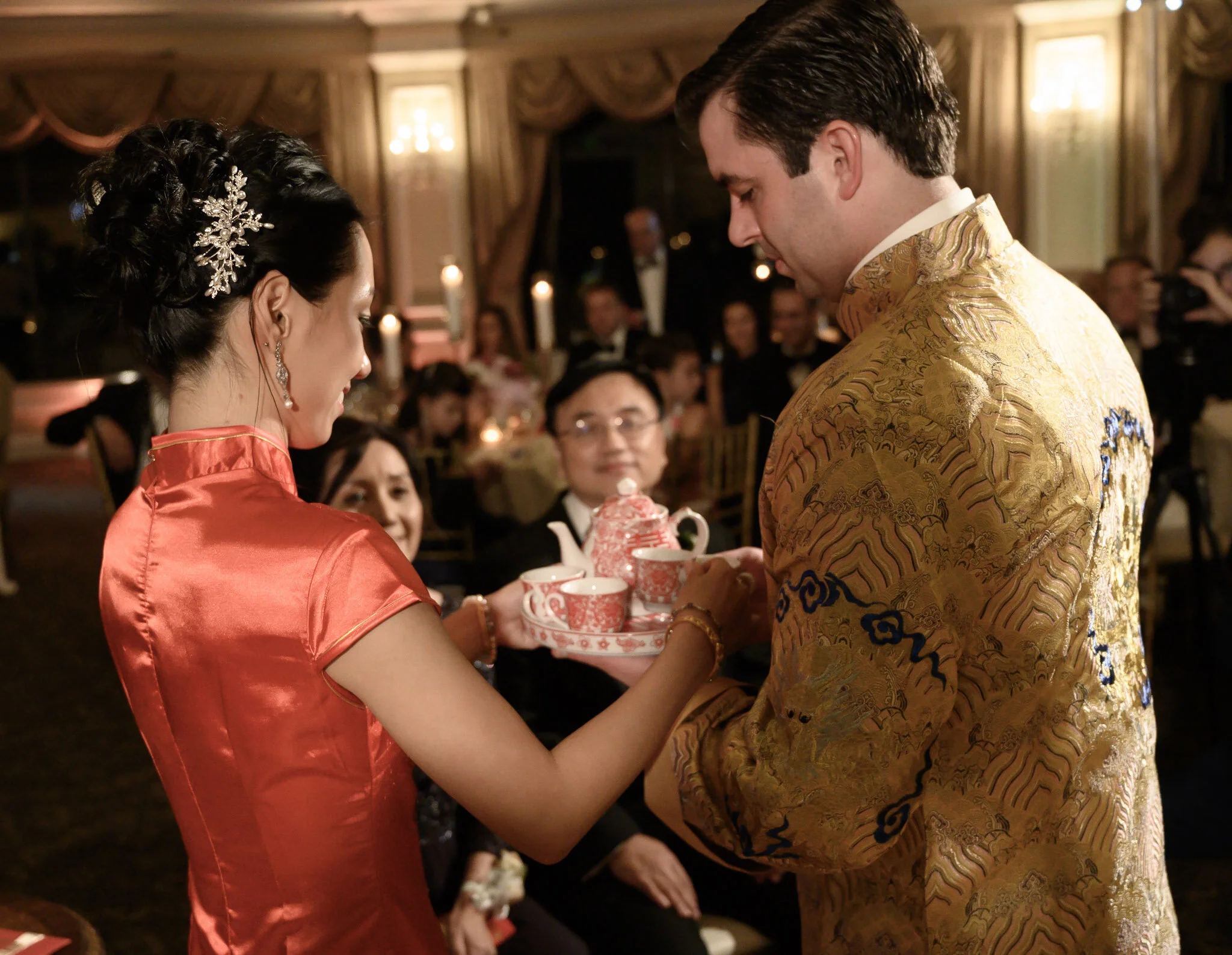 Man and woman exchanging traditional tea cups at a wedding ceremony, with guests in the background.